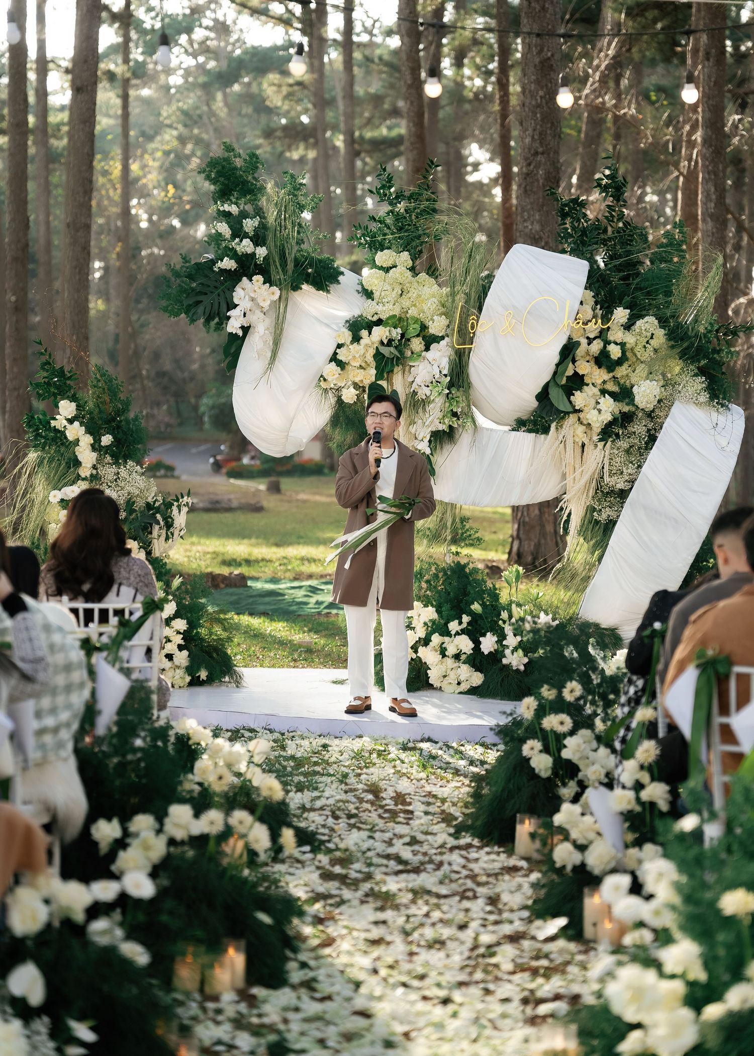 Officiant speaking at outdoor forest wedding ceremony with white floral arch.