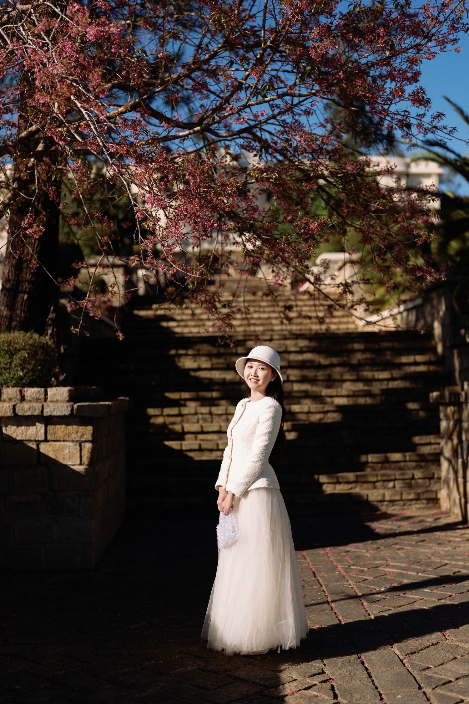Woman in white dress and hat poses near blooming pink cherry blossom tree on brick steps outdoors.