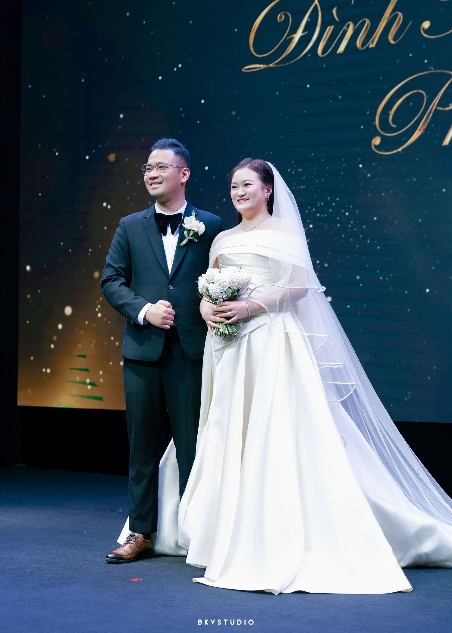 Bride in white gown holding bouquet stands with groom in black tuxedo at elegant wedding reception.