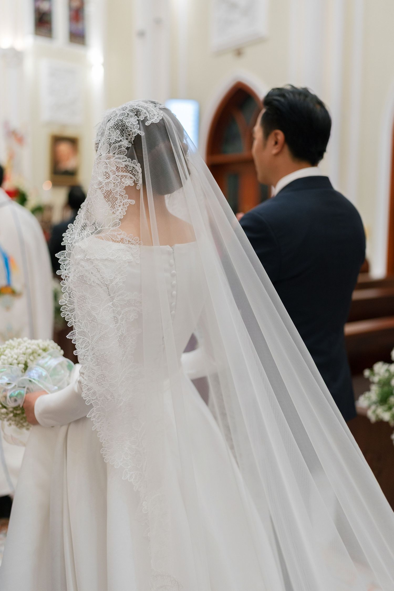 Bride in white lace gown and veil holding bouquet stands with groom in navy suit at church altar.