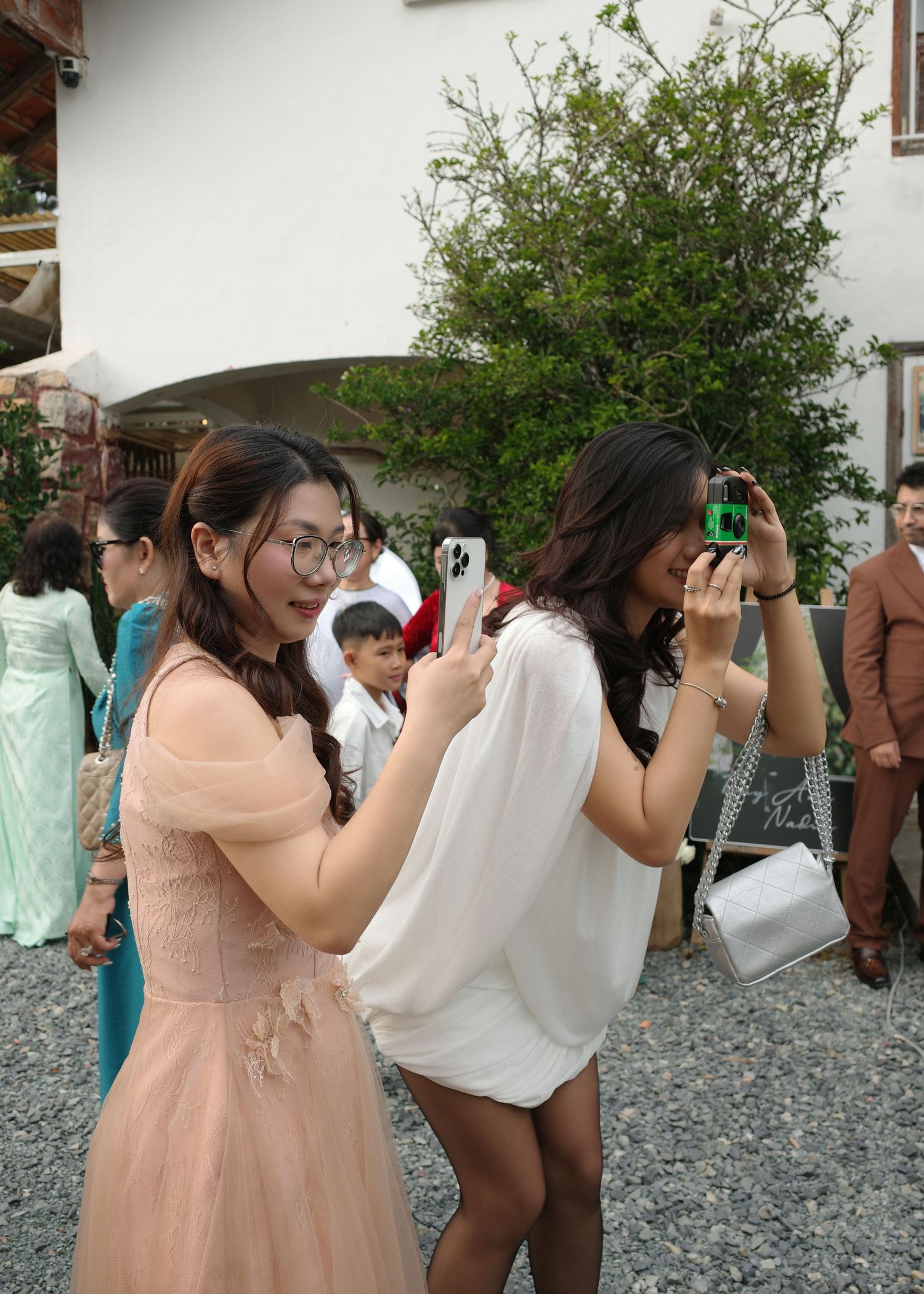 Two young women in elegant dresses photographing at an outdoor wedding celebration.