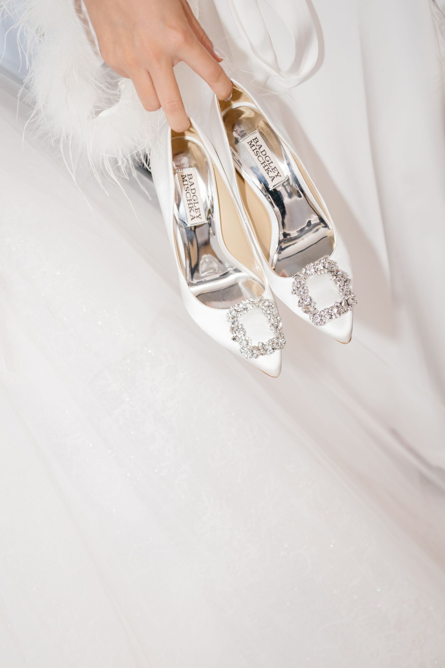 Bride holding white Badgley Mischka satin pointed-toe heels with crystal buckles against a lace wedding gown.