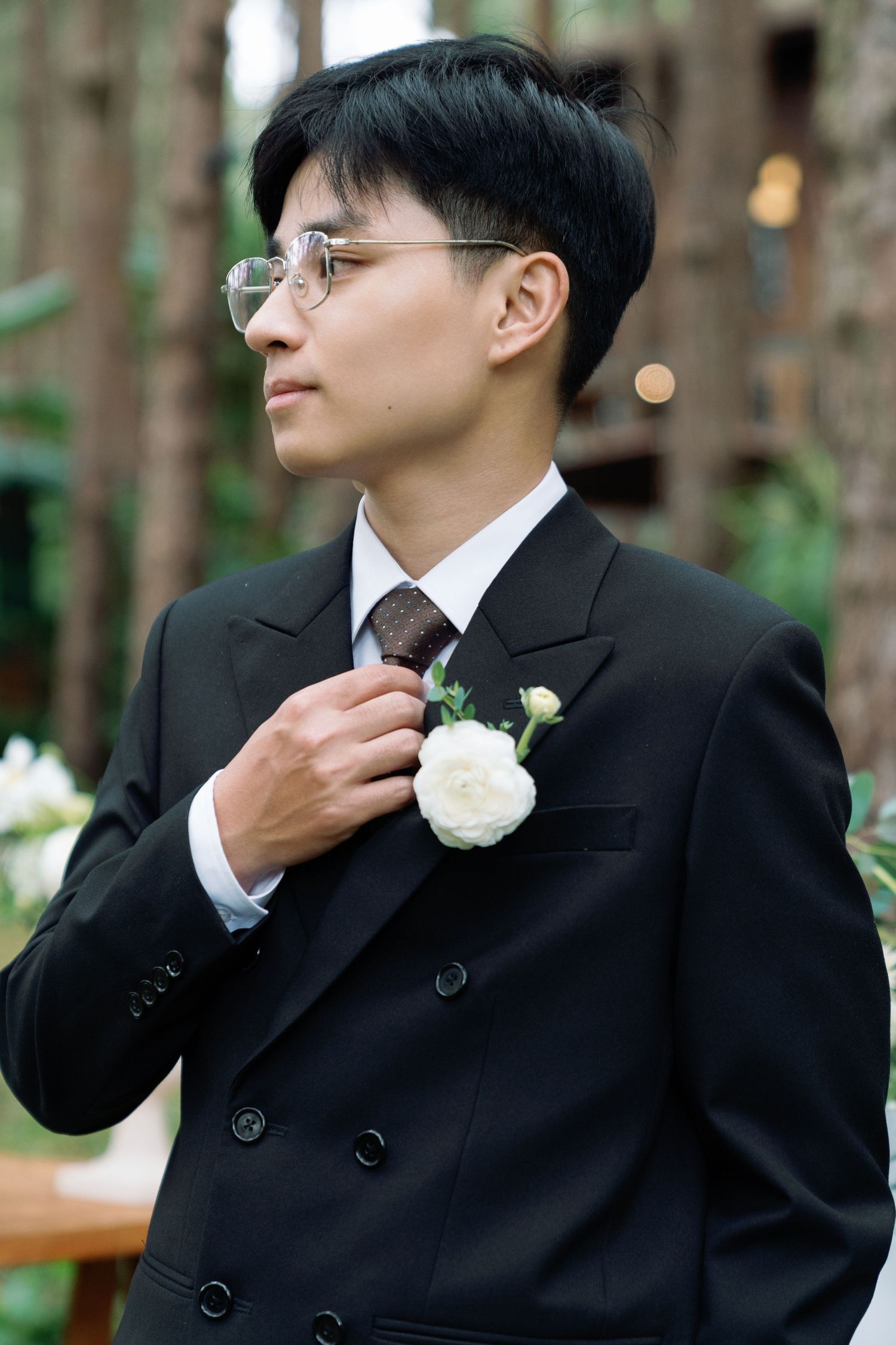 Elegant groom in black suit adjusting boutonniere with white rose outdoors at a forest wedding venue.