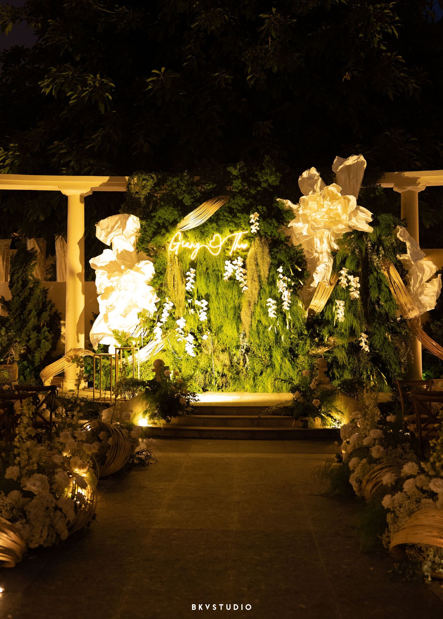 Elegant outdoor wedding ceremony aisle illuminated at night with lush greenery, white floral decor, and glowing neon signage.