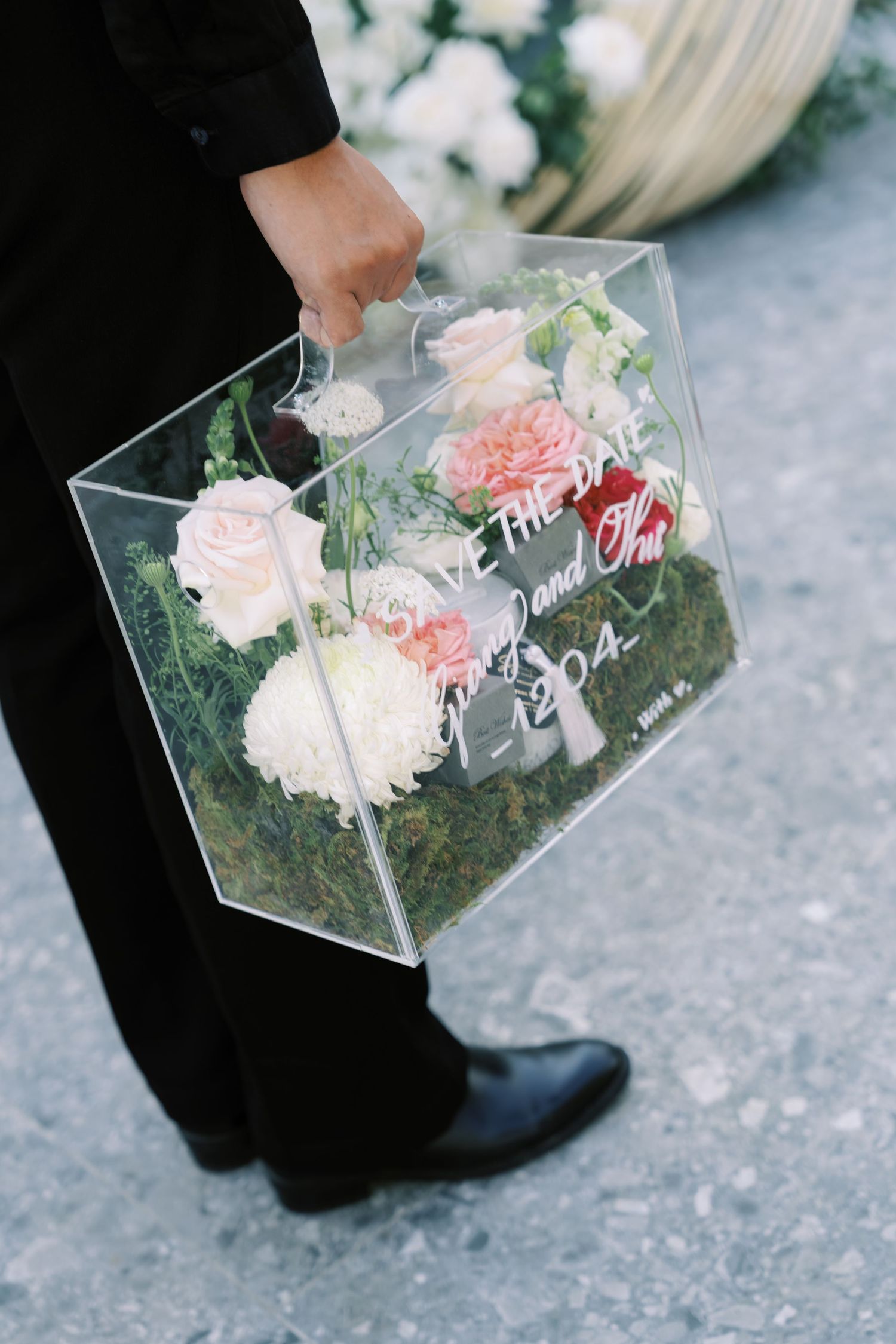 Groom holds clear acrylic box filled with pink and white roses and save the date card.
