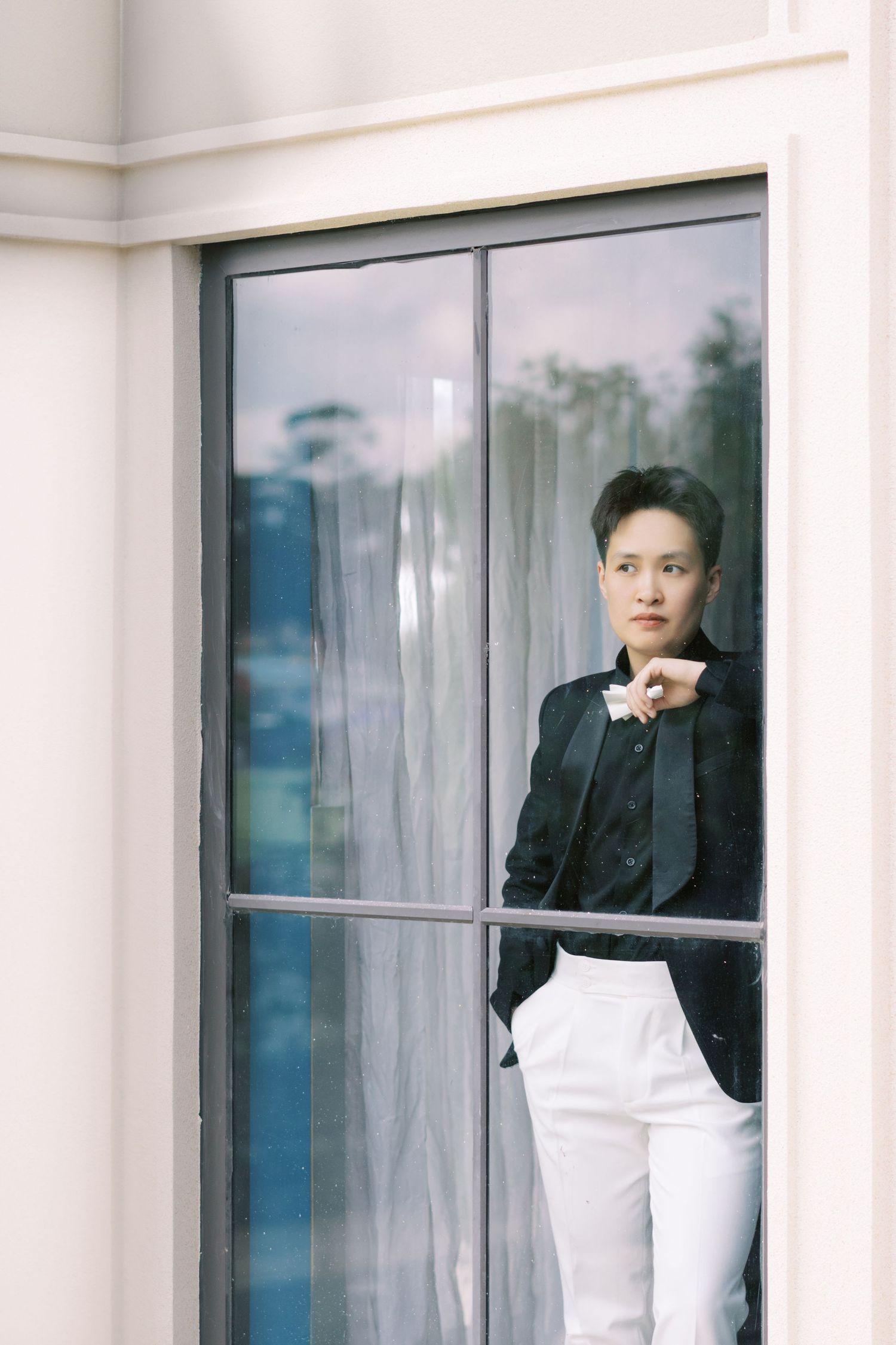 Stylish person in black blazer and white pants standing confidently by large glass window outdoors.