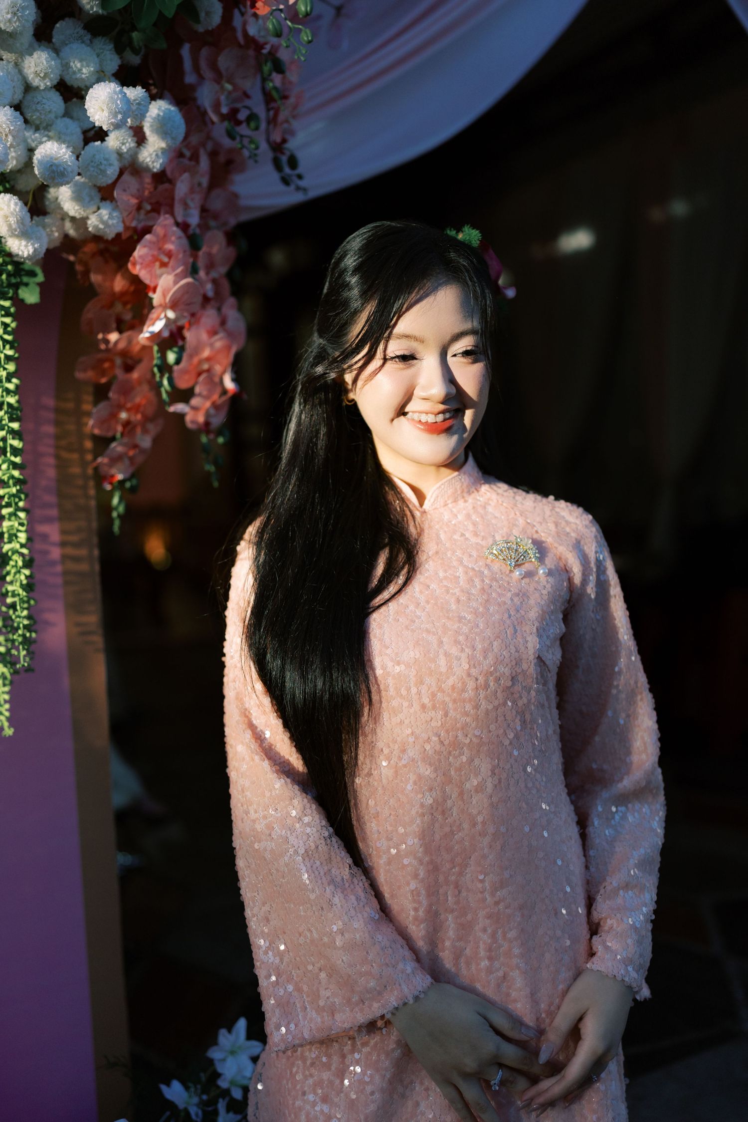 Young Vietnamese woman smiling in pink sequined dress at traditional wedding ceremony with floral decorations.