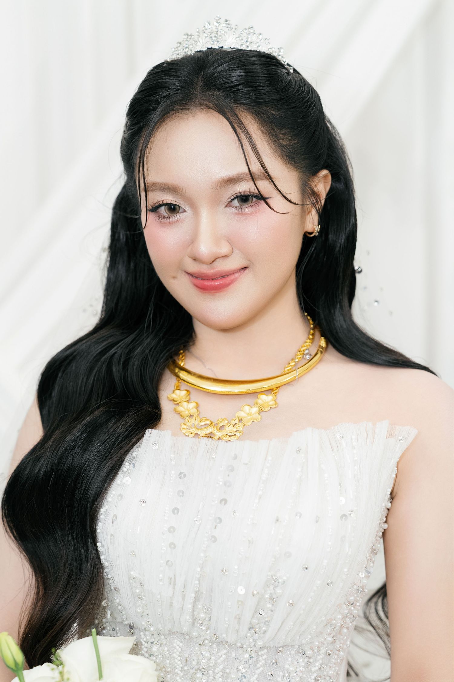 Beautiful Vietnamese bride wearing a white gown, gold necklace, and tiara with long black hair.