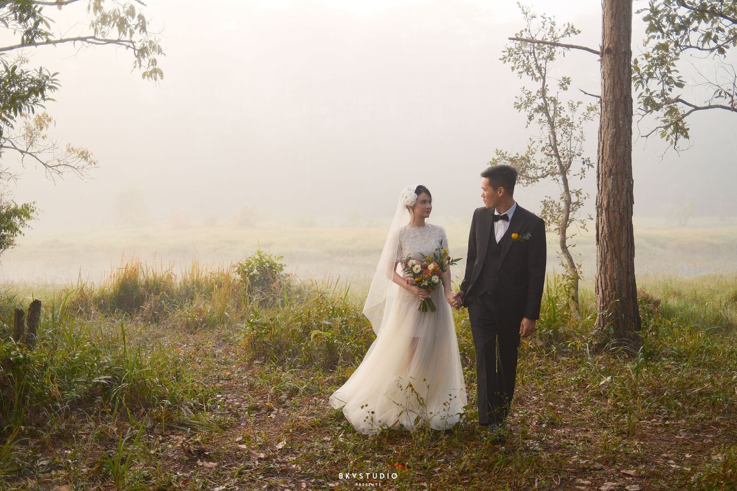 Couple in wedding attire holding hands in misty forest clearing, bride in white gown, groom in black tuxedo.