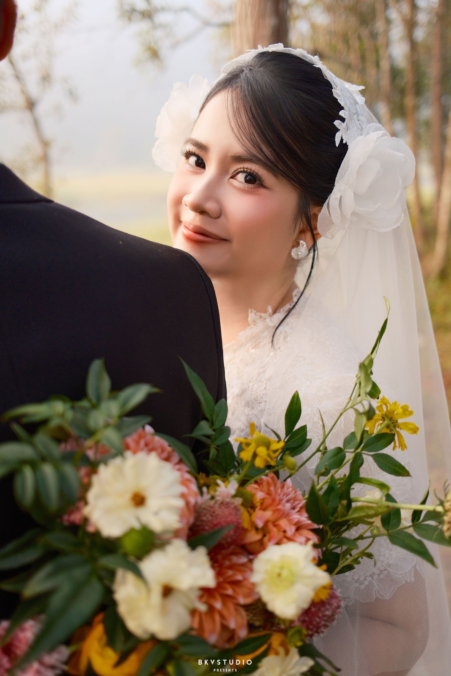 A smiling bride in a white wedding dress and veil holds a colorful bouquet of flowers outdoors.