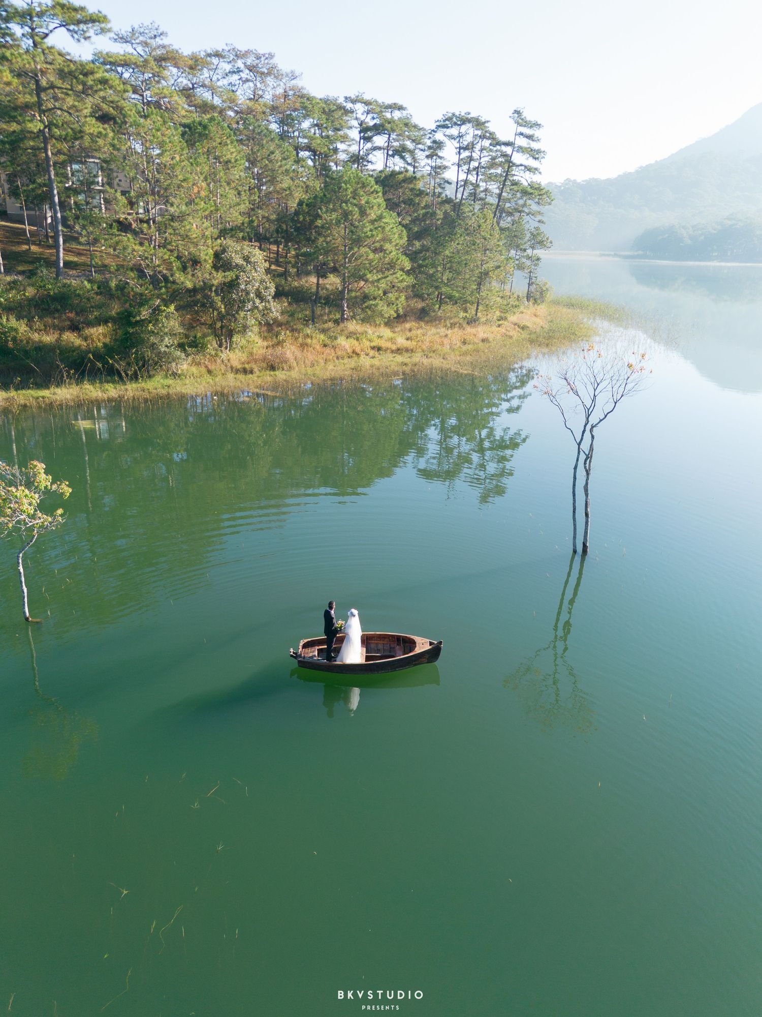Aerial view of bride and groom in small boat on calm green lake surrounded by pine trees.