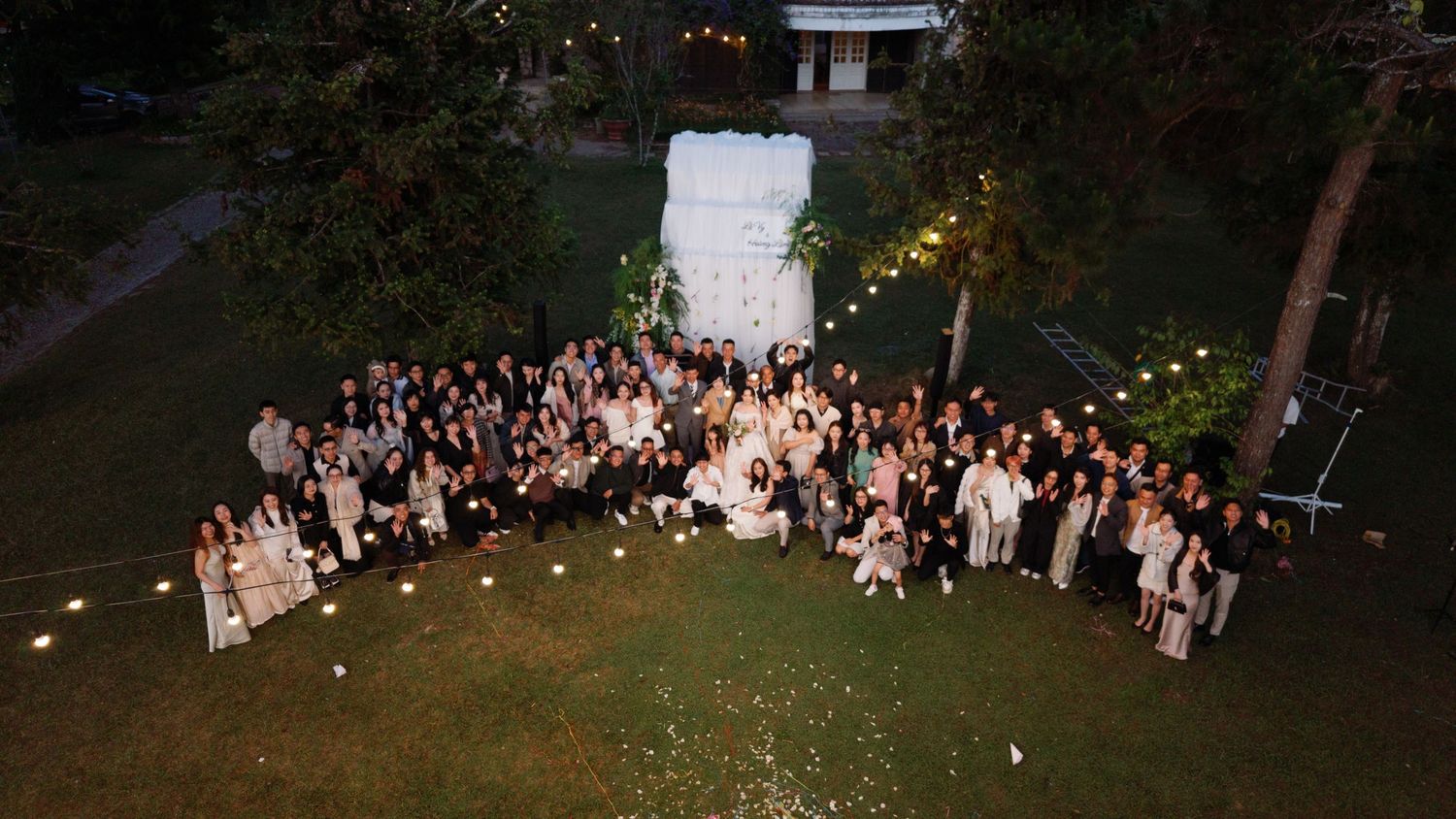 Aerial night view of wedding group photo on a lawn with string lights and white floral backdrop.