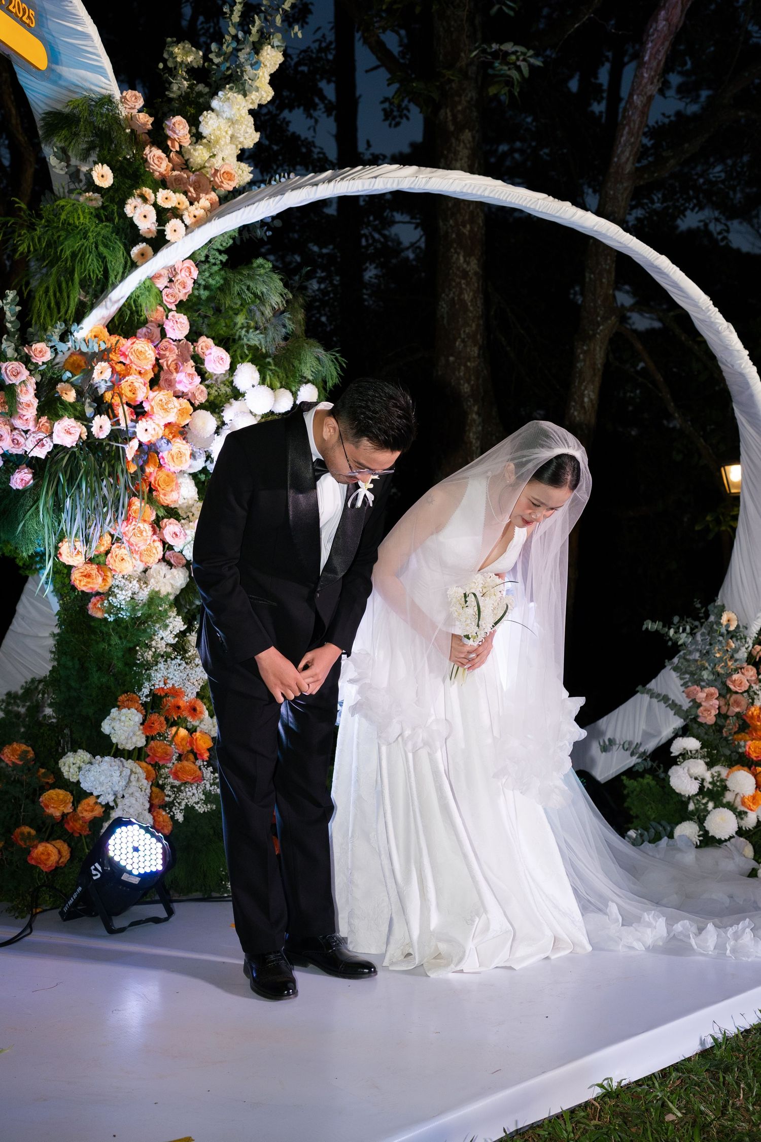Bride and groom bowing at floral circular arch decorated with orange and white flowers at outdoor wedding.