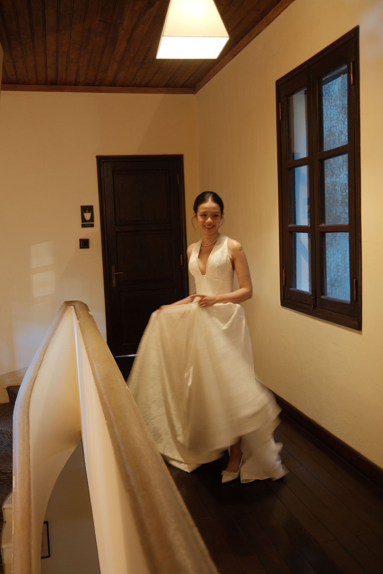 A radiant bride in a deep V-neck white wedding gown walks along a warmly lit indoor hallway.
