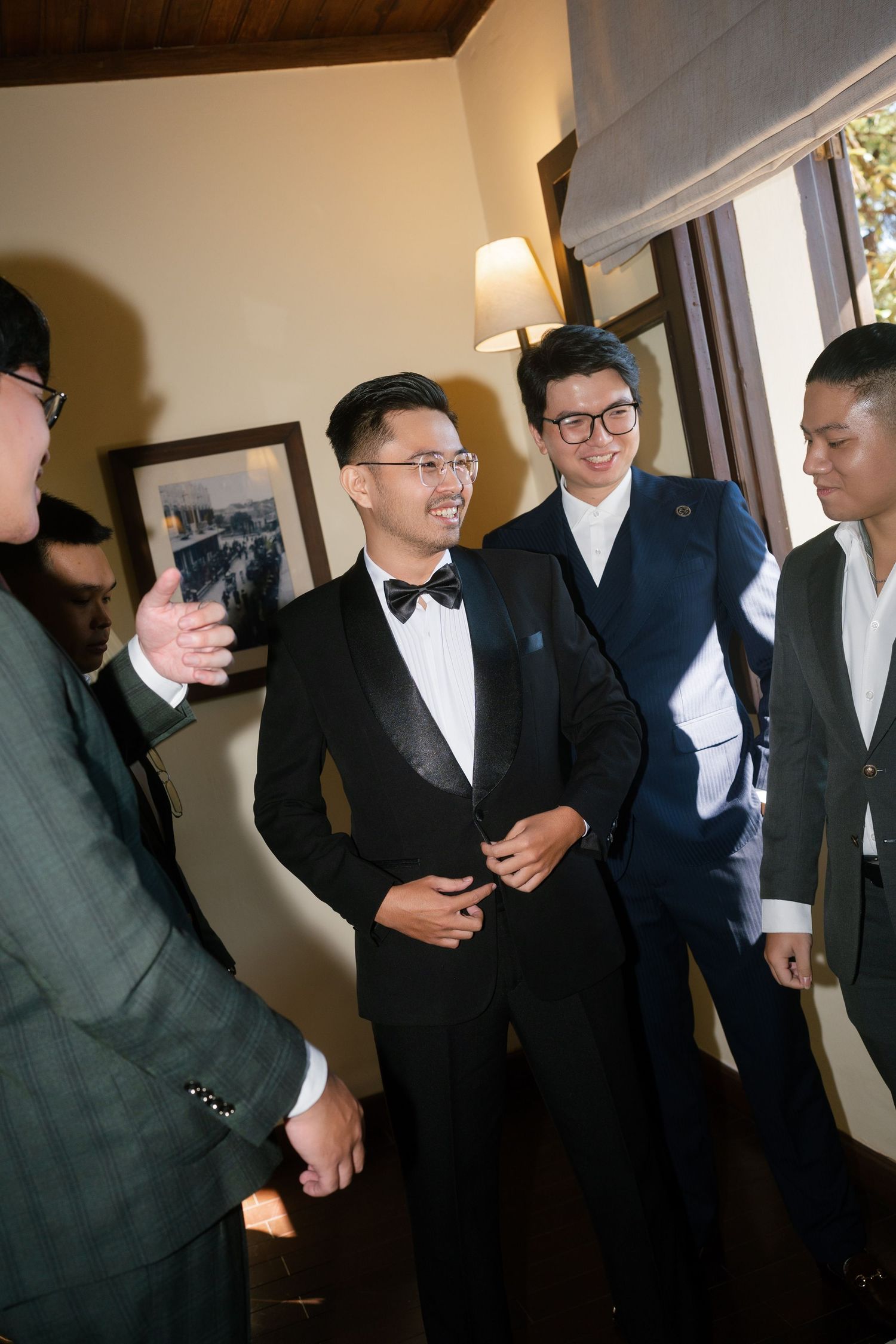 Groom in black tuxedo laughing with groomsmen in suits indoors before wedding ceremony.
