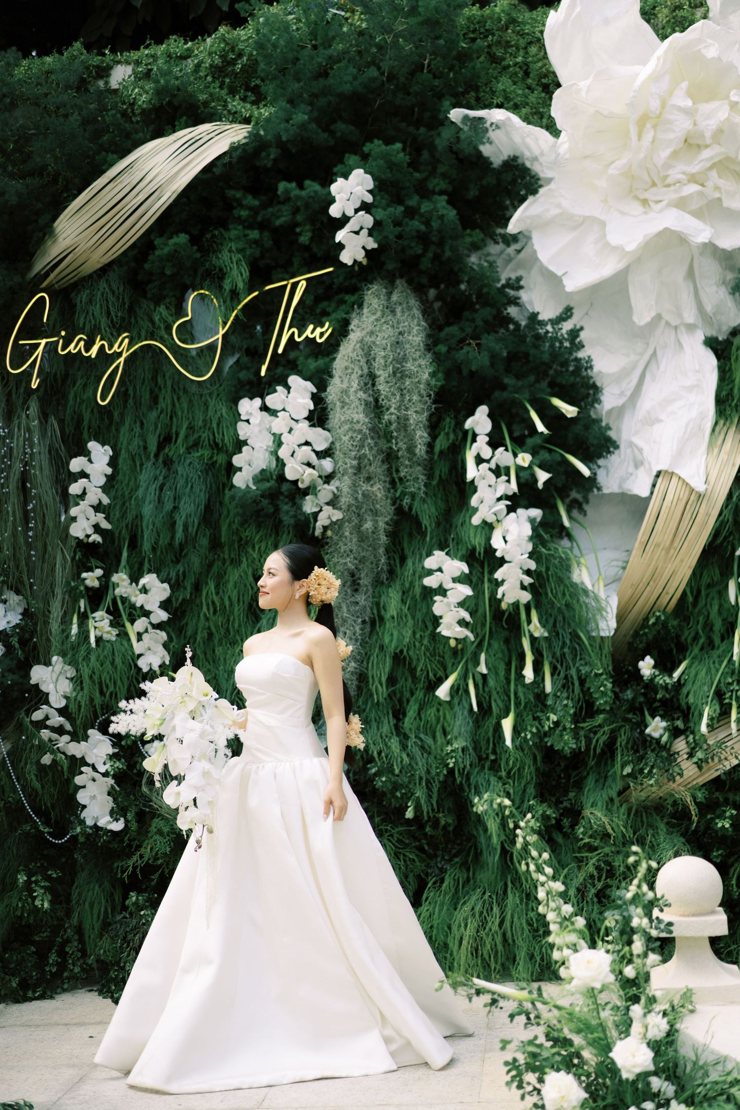 Bride in white gown poses before lush green floral backdrop with large white blooms at wedding of Giang and Thu.