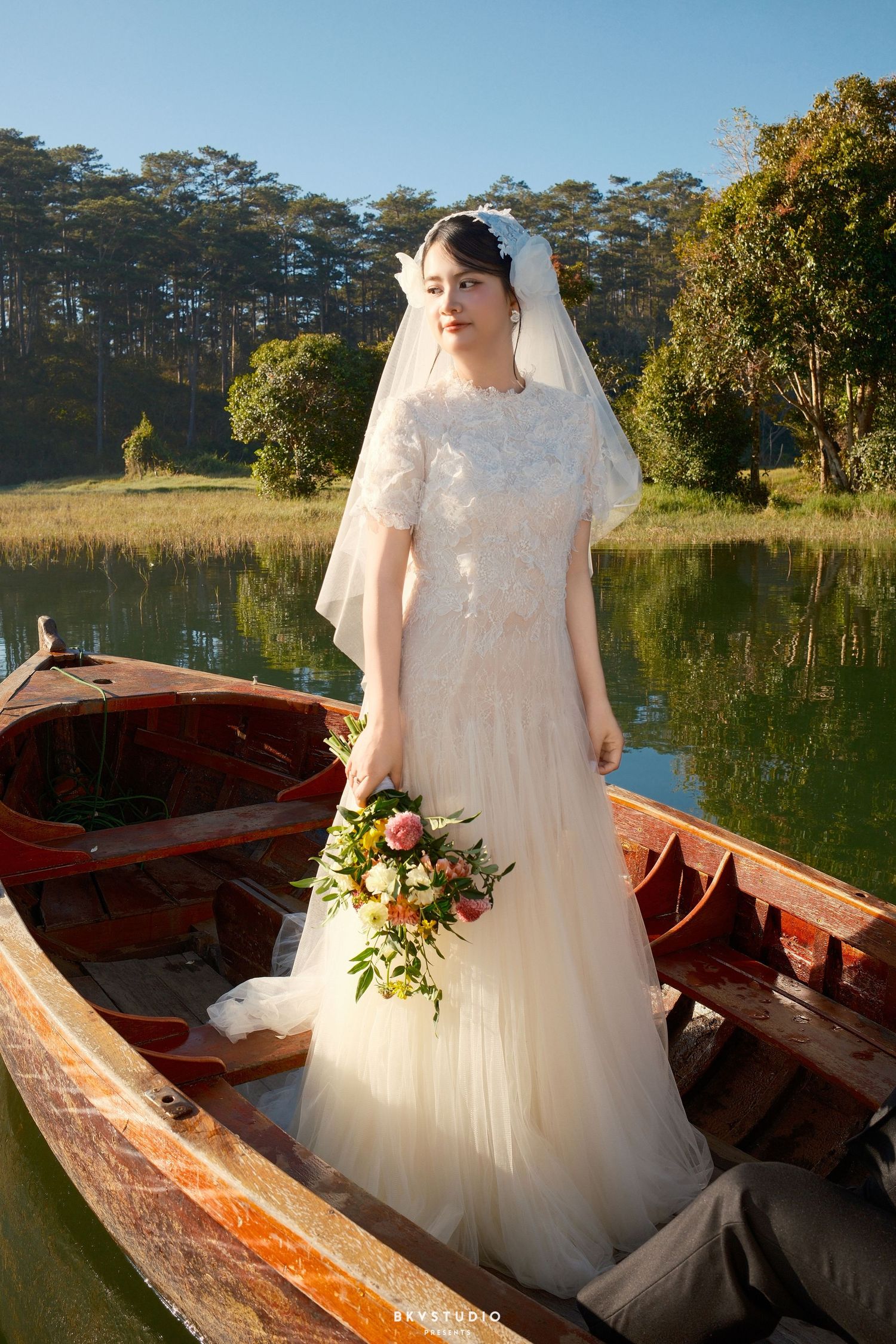 Beautiful bride in white lace dress and veil stands in a rustic wooden boat holding a floral bouquet.