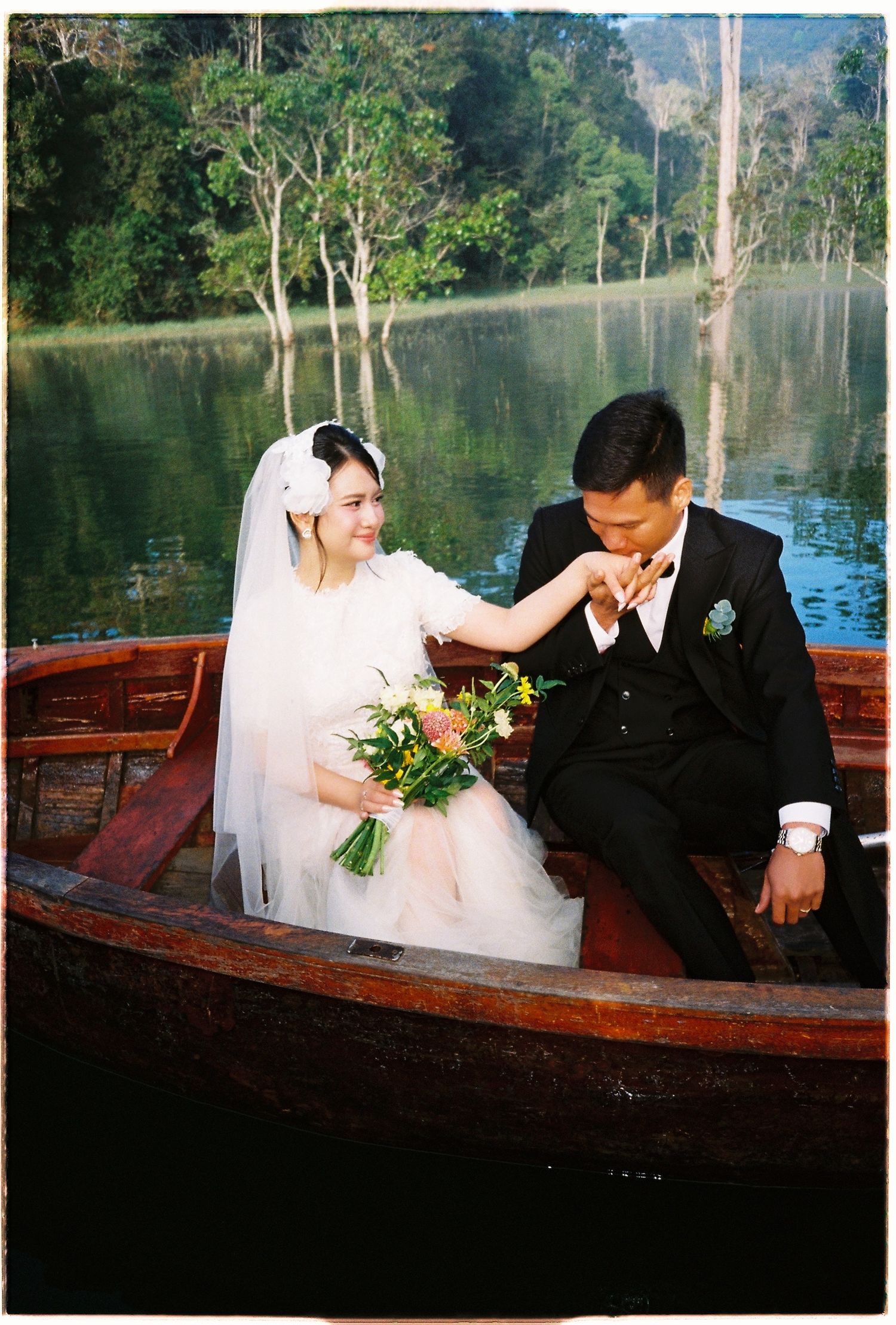 Groom kisses bride's hand as she holds a floral bouquet while seated in a wooden boat on a lake.