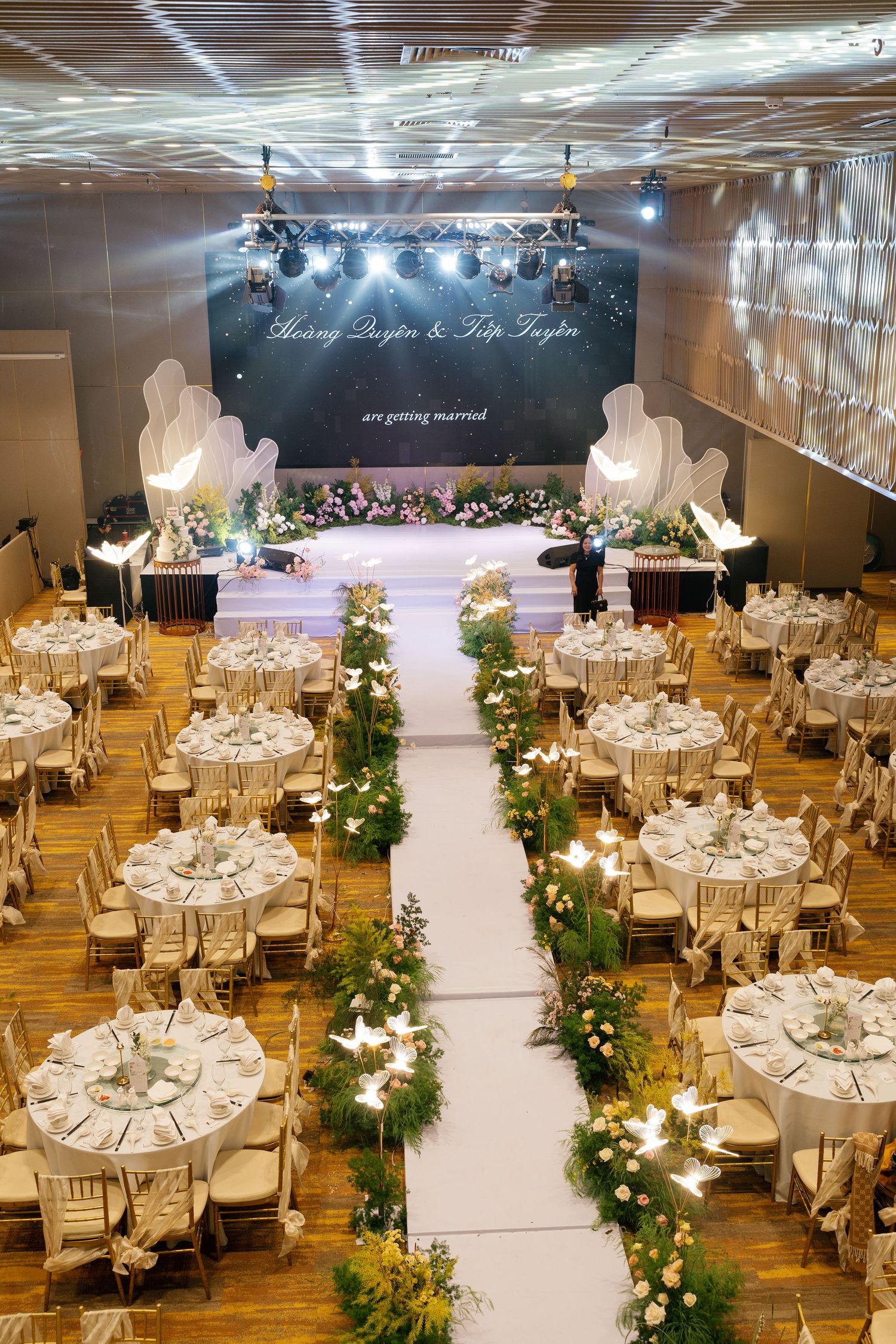 Elegant wedding reception hall with floral aisle, round tables, and illuminated stage backdrop.