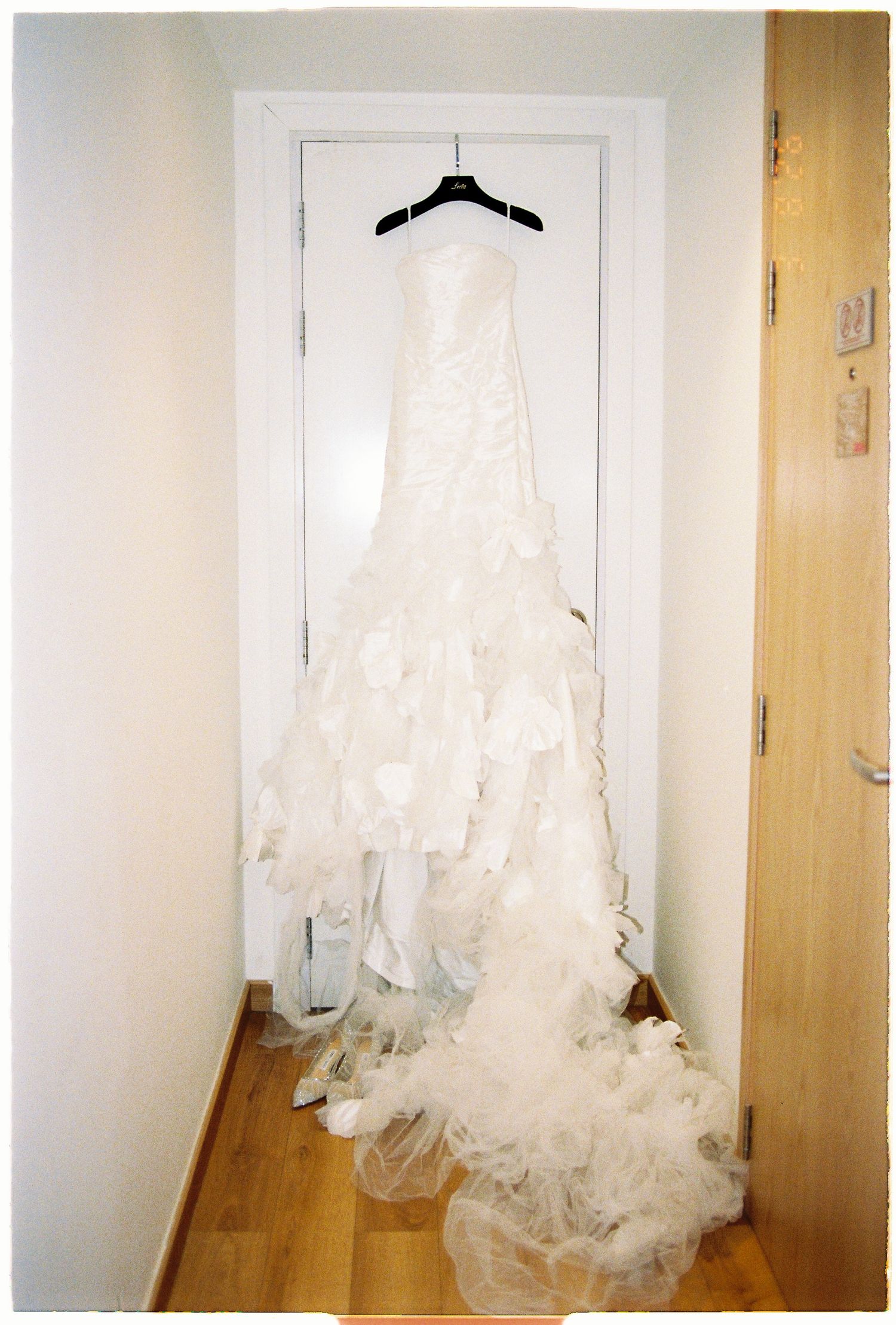 Elegant white wedding dress with ruffled skirt displayed on hanger against white door in hallway.