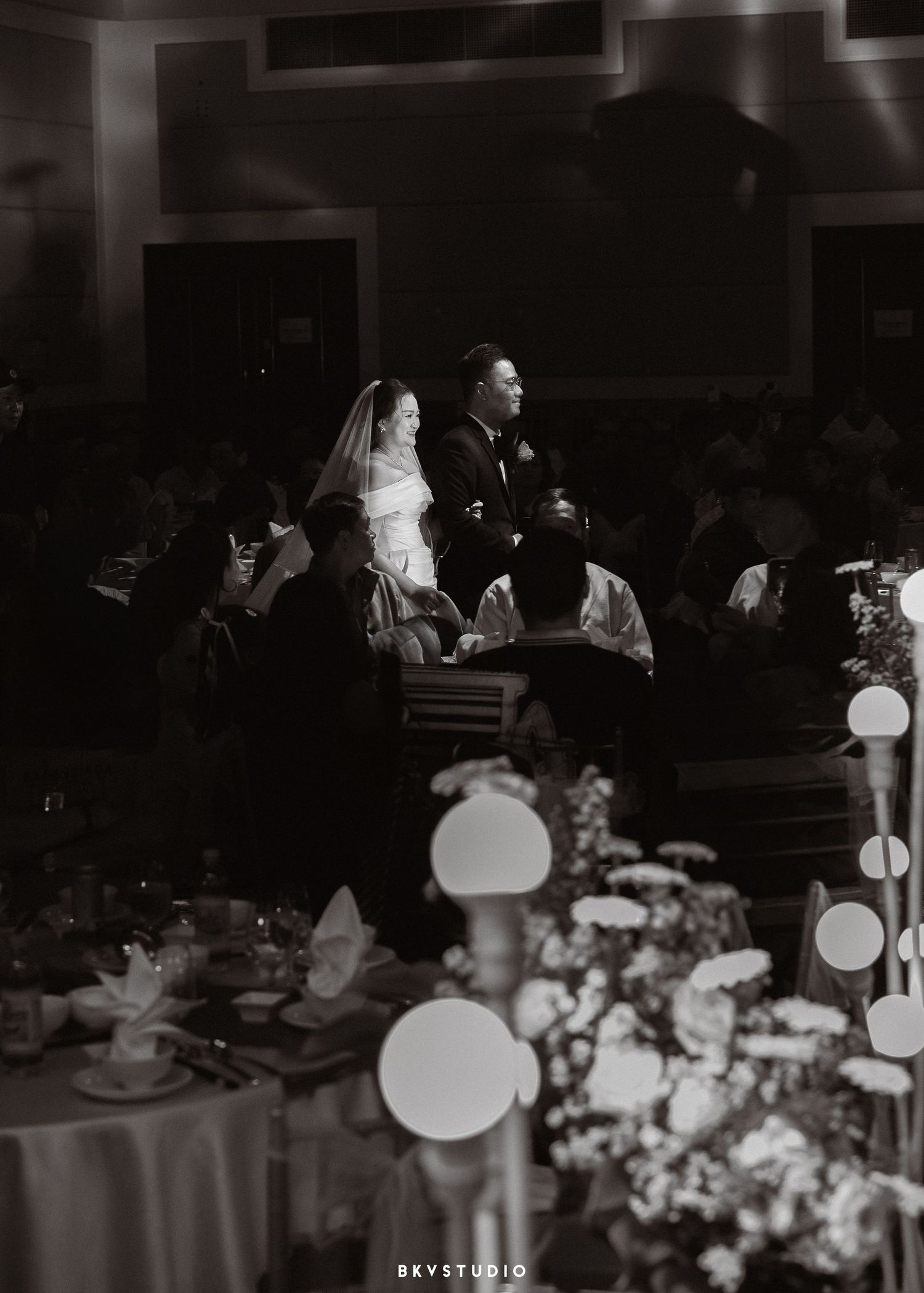 Black and white wedding reception photo of bride and groom seated at head table, surrounded by guests and floral arrangements.