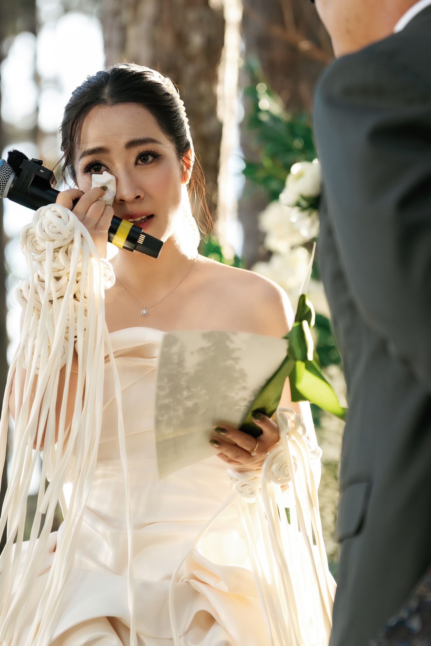 Emotional bride in white strapless gown wiping tears during outdoor wedding ceremony while holding bouquet.