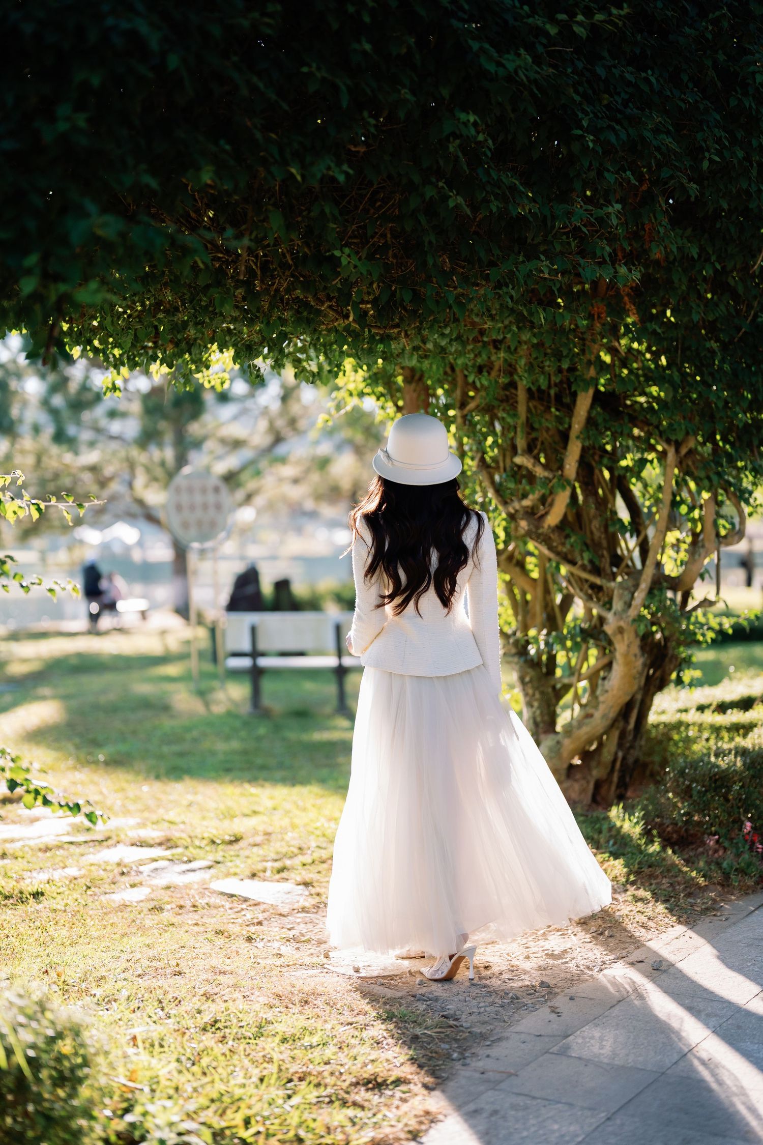 Woman in white dress and hat walks along a sunny park path beside lush green trees.