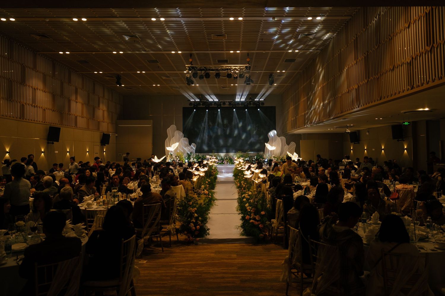 Elegant wedding reception hall with floral aisle, candlelit tables, and glowing stage backdrop filled with seated guests.