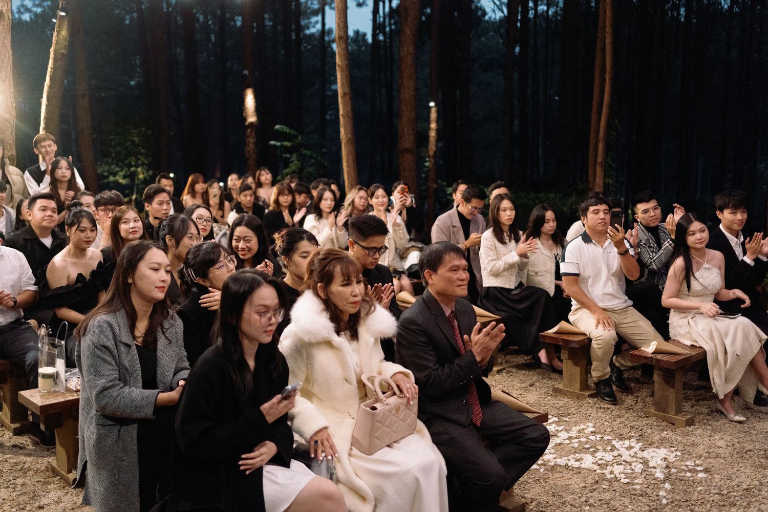 Wedding guests seated outdoors in a forest venue at dusk, applauding during a romantic ceremony.