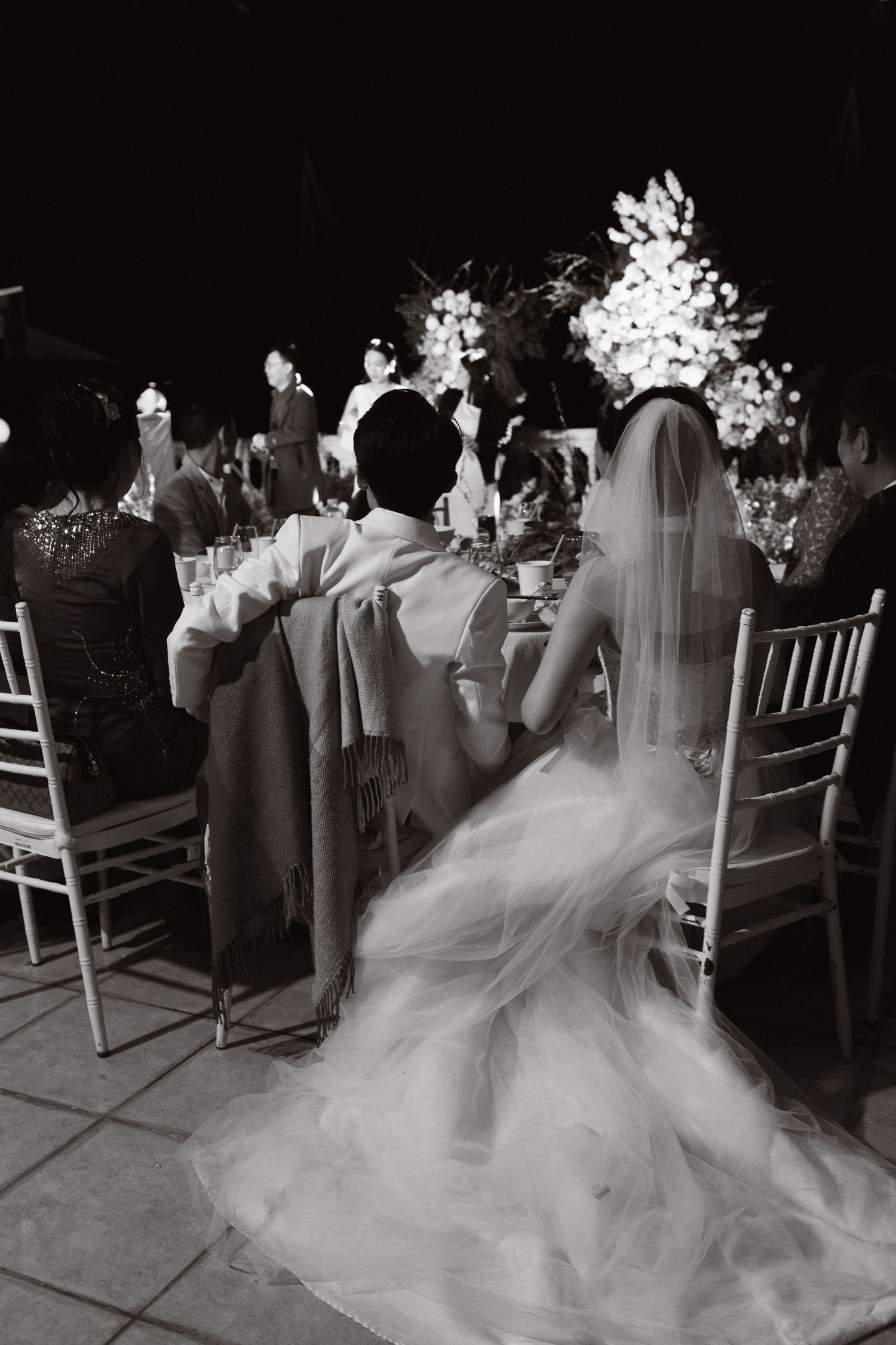 Black and white photo of bride and groom seated at wedding reception table, bride's veil cascading elegantly.