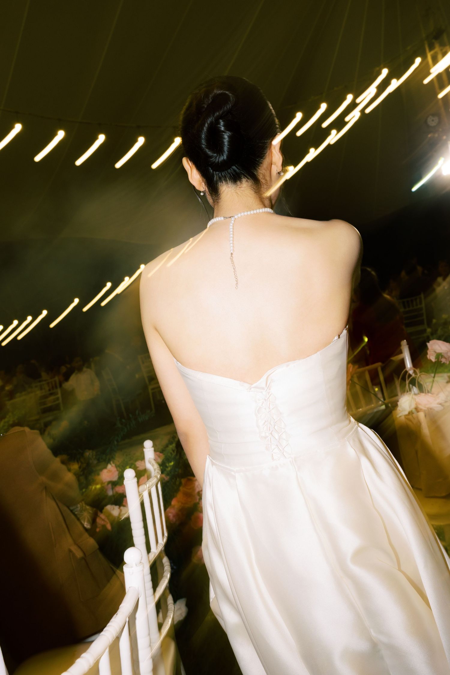 Bride in white halter wedding gown viewed from behind at elegant reception venue with warm lighting.