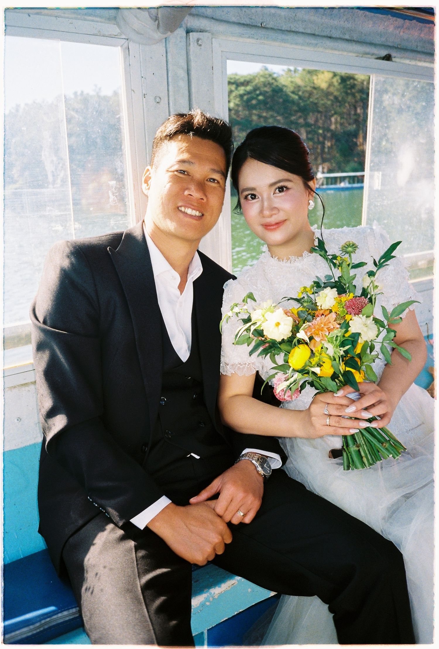 A smiling couple in wedding attire sits together inside a boat, bride holding a colorful bouquet.