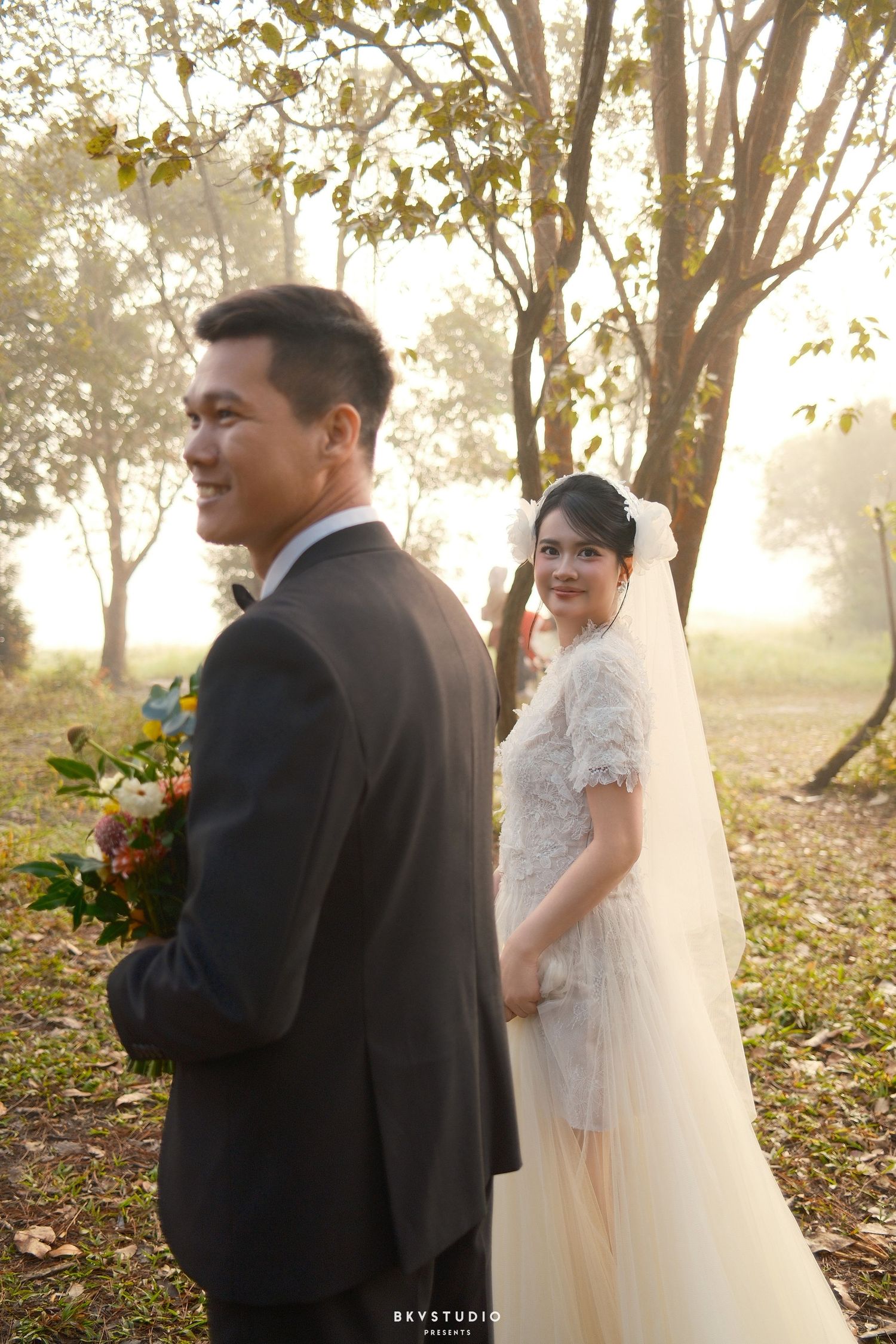 Bride and groom stand back-to-back in misty forest, both smiling, groom holding colorful bouquet.