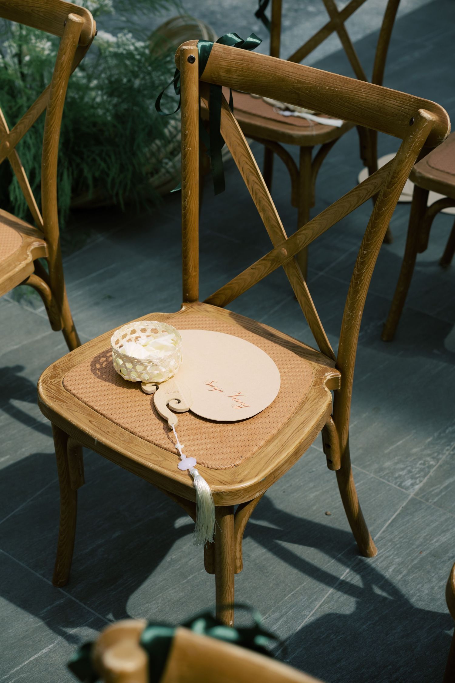 Wooden cross-back chair with a round fan program and small decorative box at an outdoor wedding ceremony.