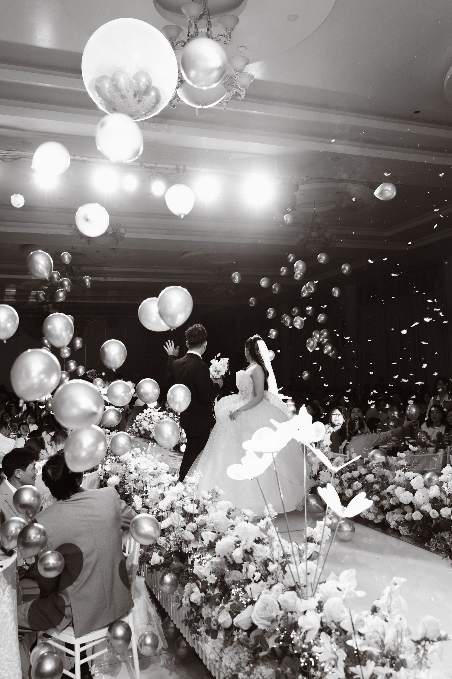 Black and white photo of bride and groom surrounded by floating balloons at grand wedding reception.