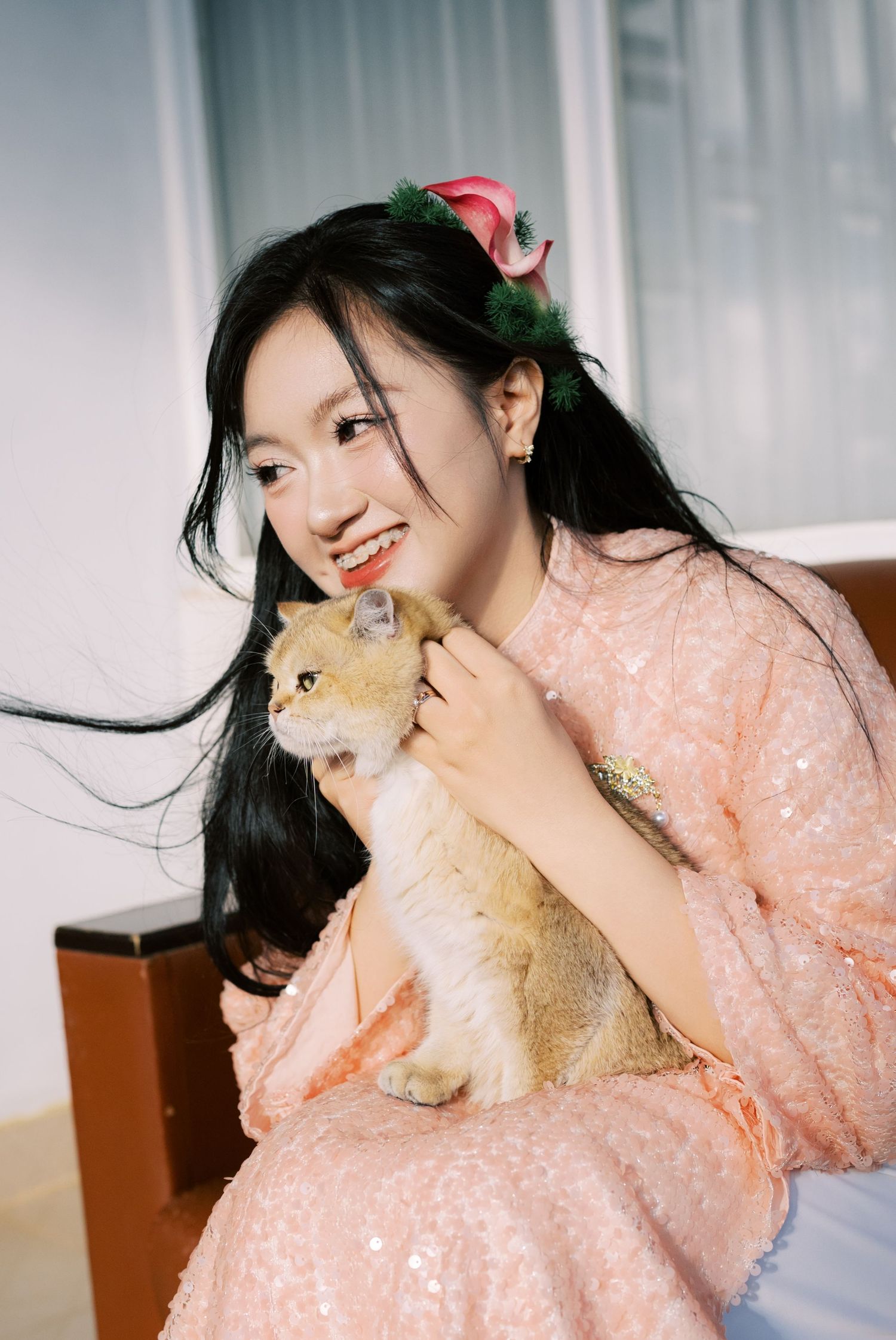 Smiling young woman in pink sequin dress holding an orange cat, wearing floral hair accessories.