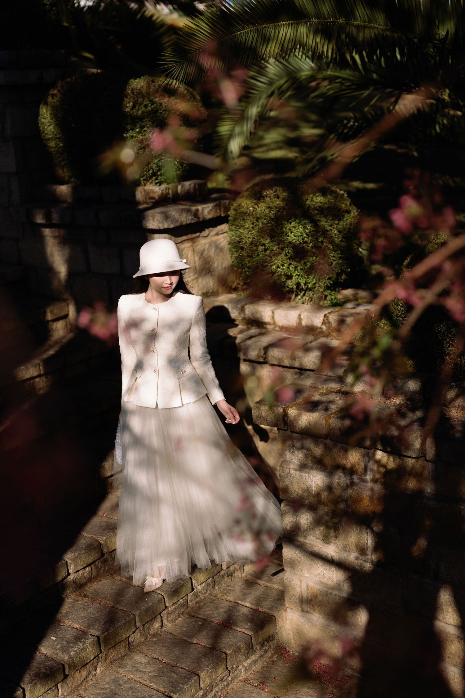 Woman in white jacket and hat standing on brick path surrounded by lush garden greenery and pink flowers.