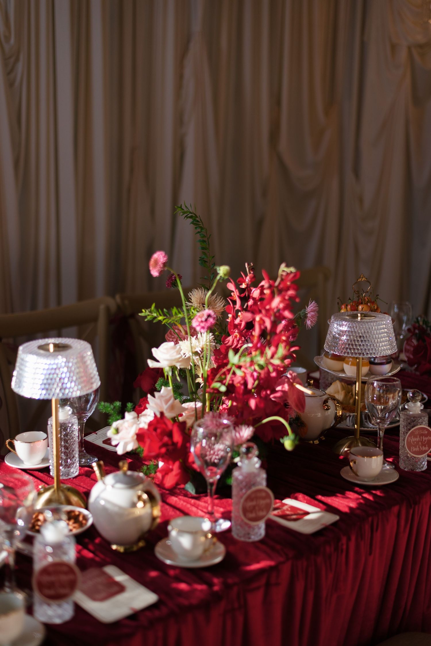 Elegant red tablecloth wedding reception table with crimson floral centerpiece, crystal lamps, white teapots, and fine china place settings.
