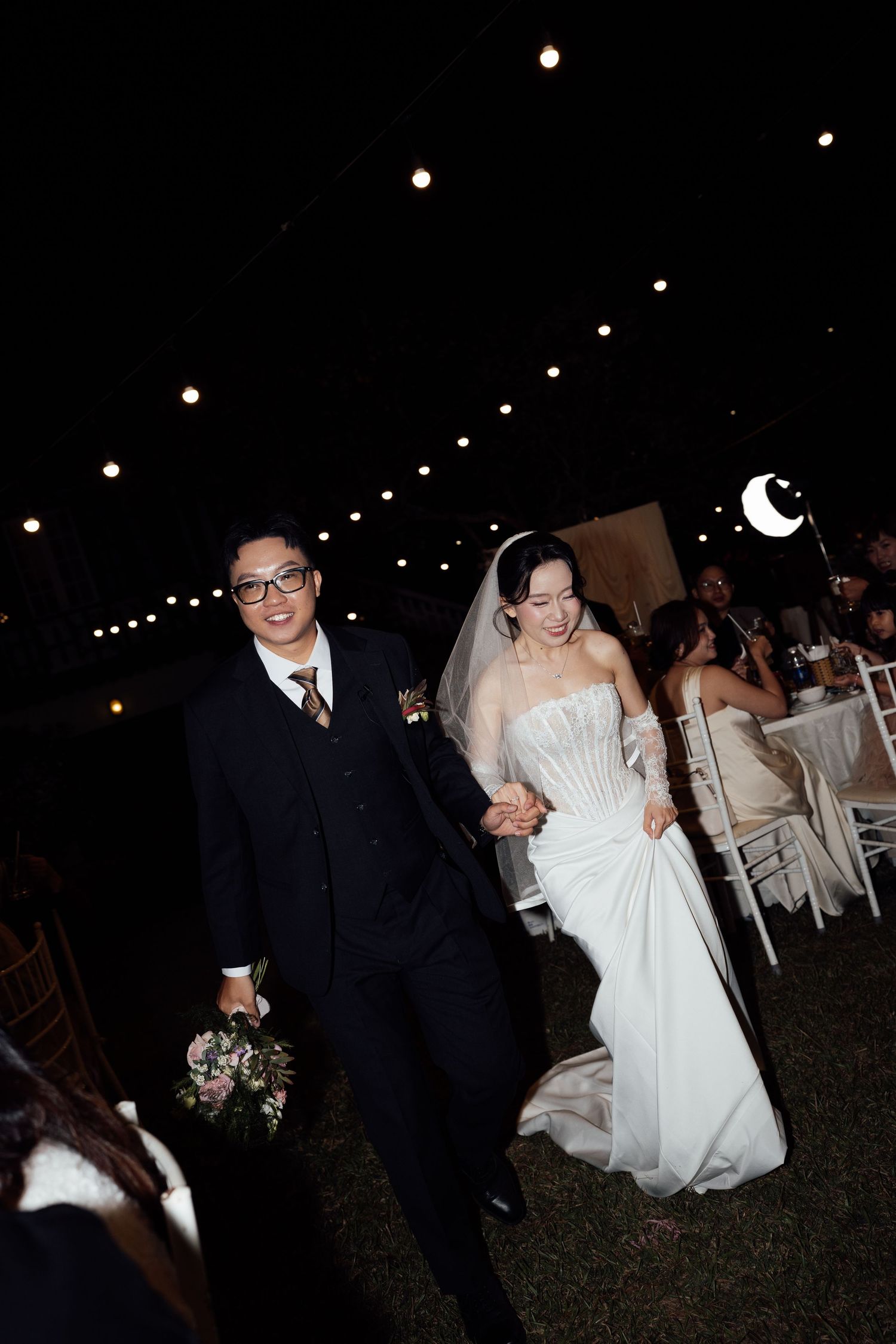Newlywed couple walking hand in hand at outdoor night wedding reception under string lights, bride in white gown and groom in dark suit.