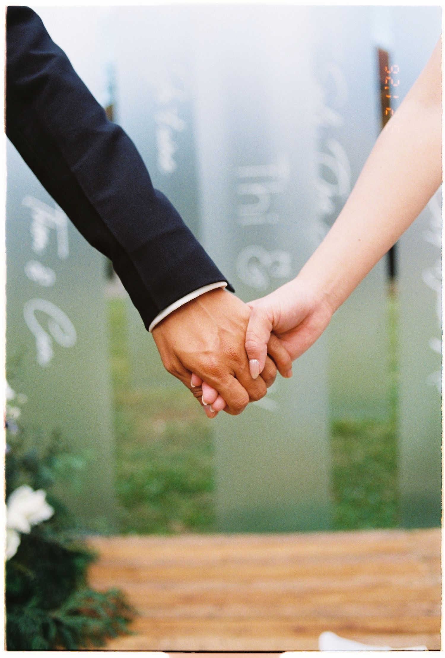 A couple holds hands at their wedding ceremony, the groom in a dark suit and bride in white, with elegant script decor behind them.