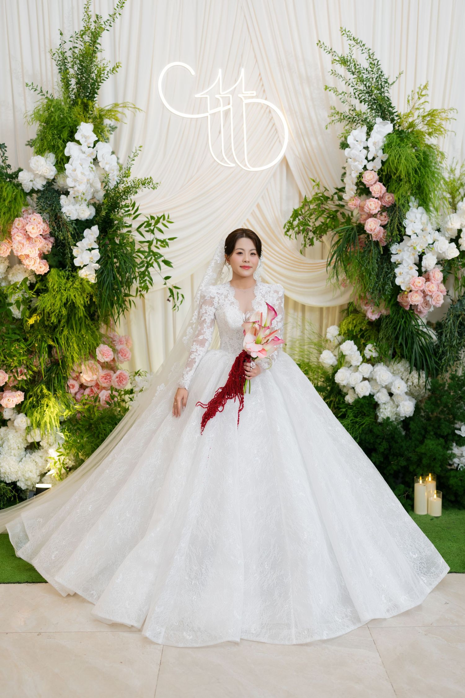 Bride in elegant white ball gown with lace sleeves holds bouquet before floral arch backdrop at wedding ceremony.