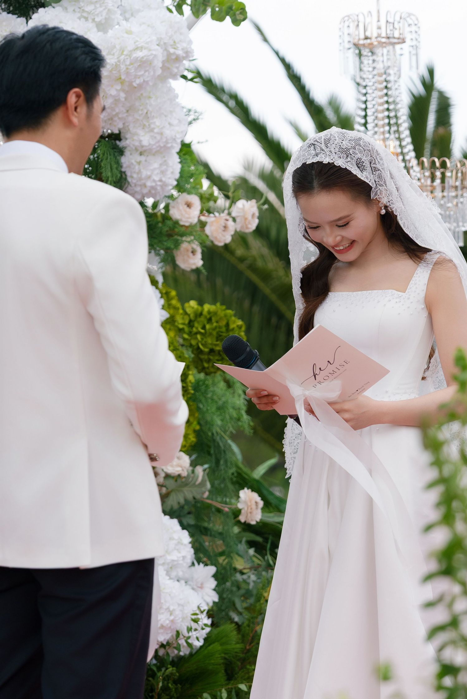 Bride in white gown and veil reads wedding vows from a pink booklet at an outdoor ceremony with floral arch.