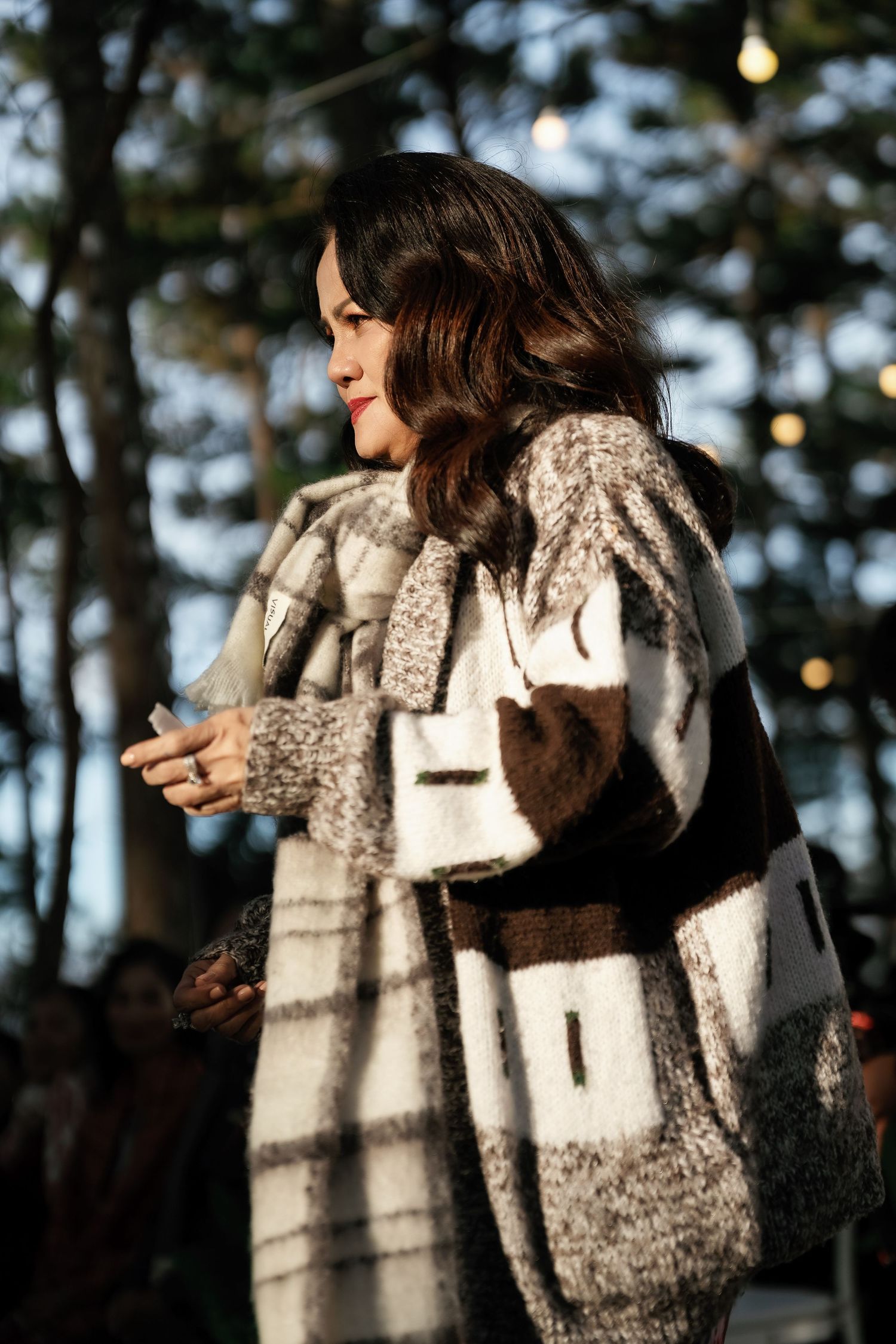 Woman in stylish plaid wool coat standing outdoors at evening event with bokeh lights.
