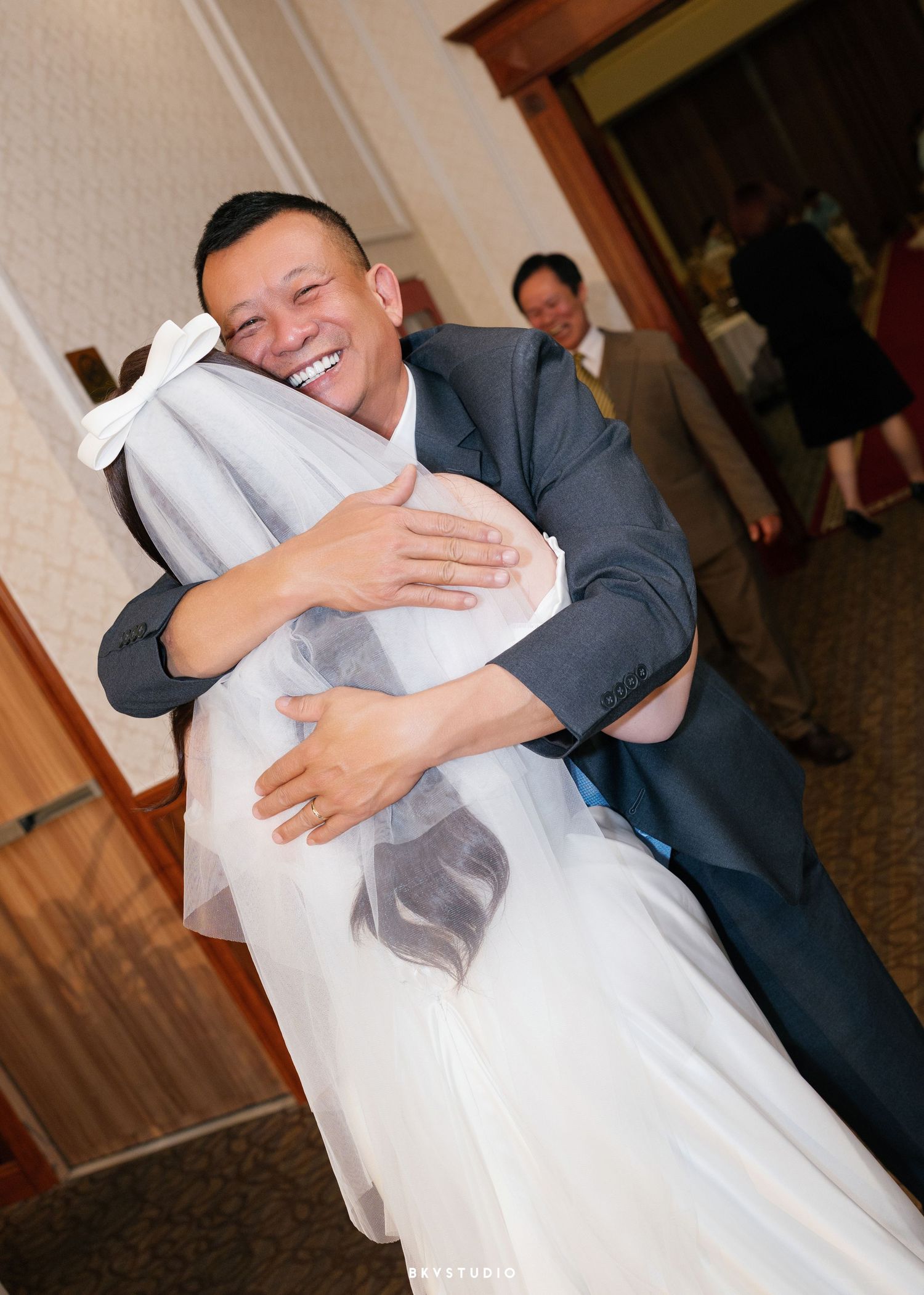 A groom in a gray suit joyfully hugs a bride in a white wedding dress and veil at a wedding reception.