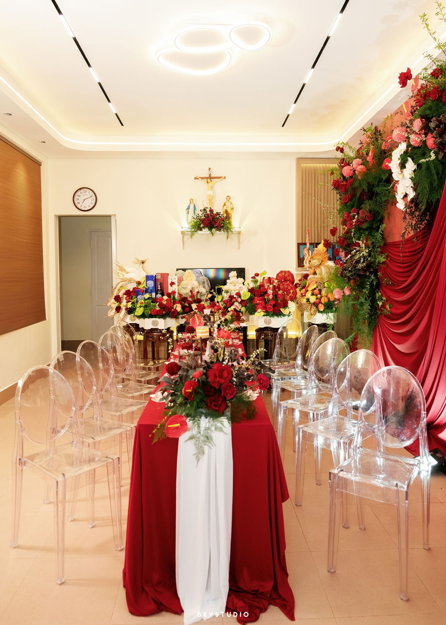 Elegant wedding reception room with red and white floral centerpieces, ghost chairs, and lush red drapery backdrop.