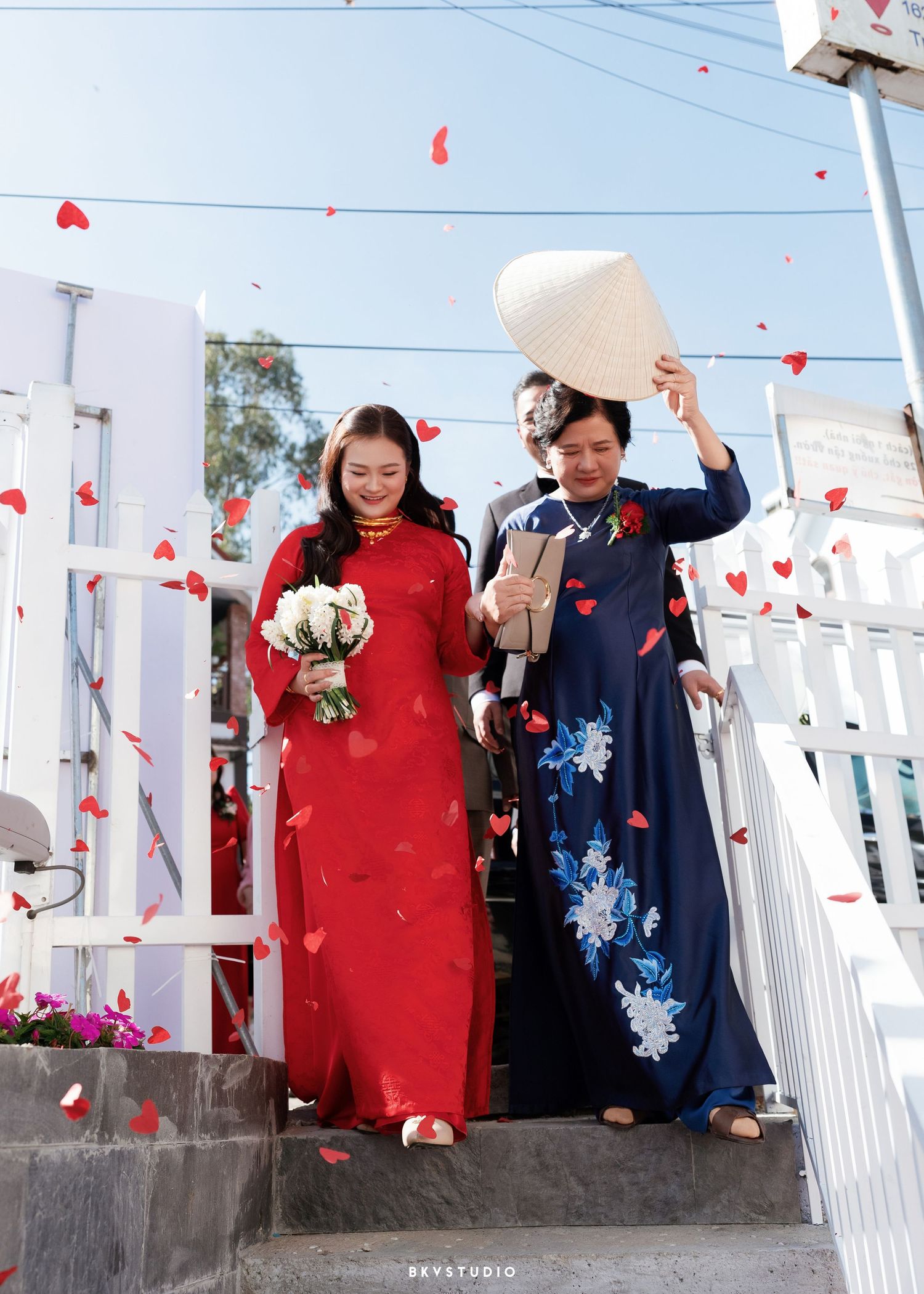 Two women in traditional Vietnamese ao dai dresses descend steps amid falling rose petals at a wedding ceremony.