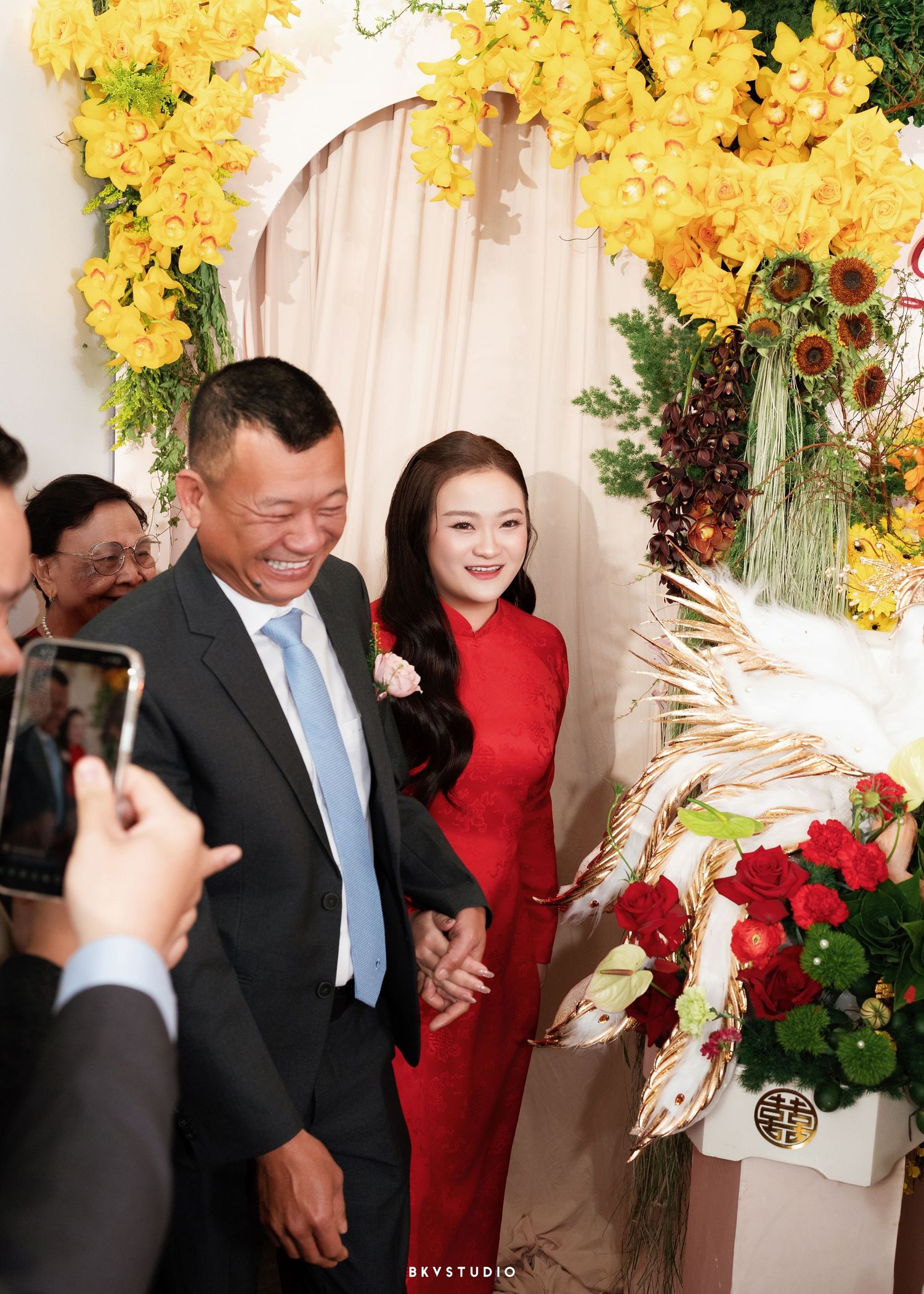 A man and woman in traditional Vietnamese wedding attire walk under yellow floral arch at a ceremony.