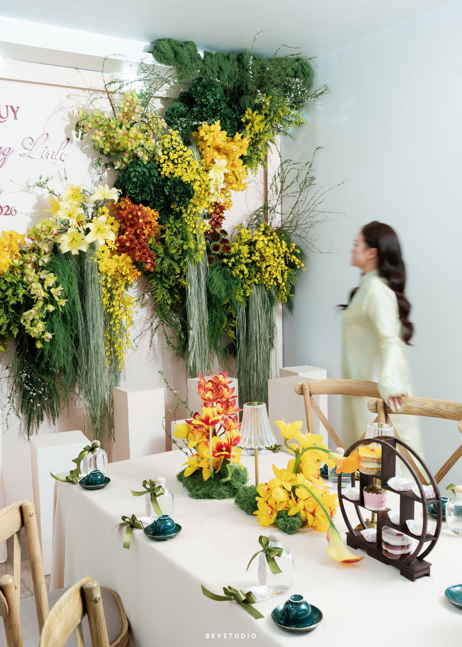 Elegant floral display with yellow orchids and greenery on a decorated table, woman in green dress in background.