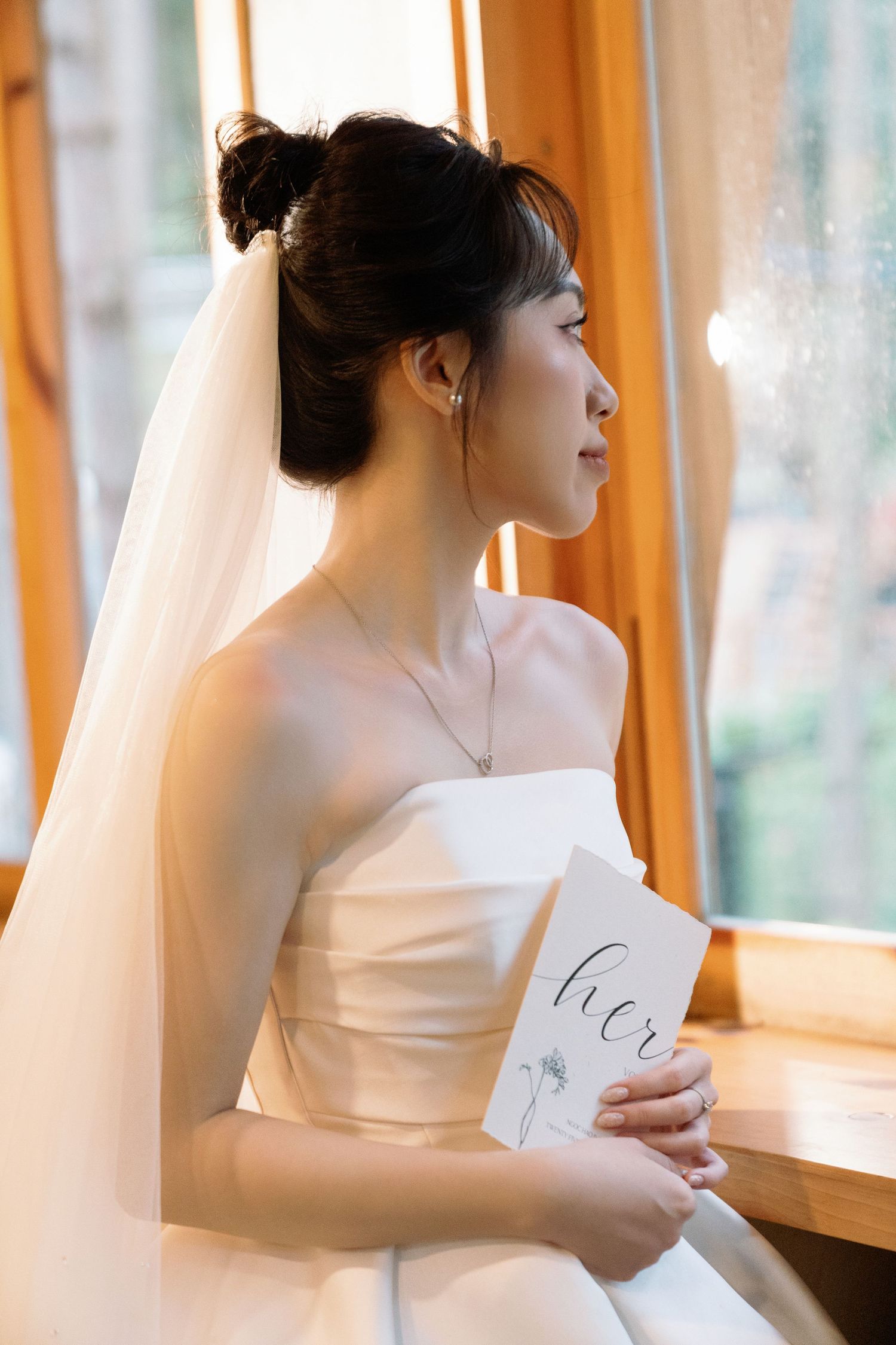 A bride in a white strapless gown and veil holds a card near a sunlit window.
