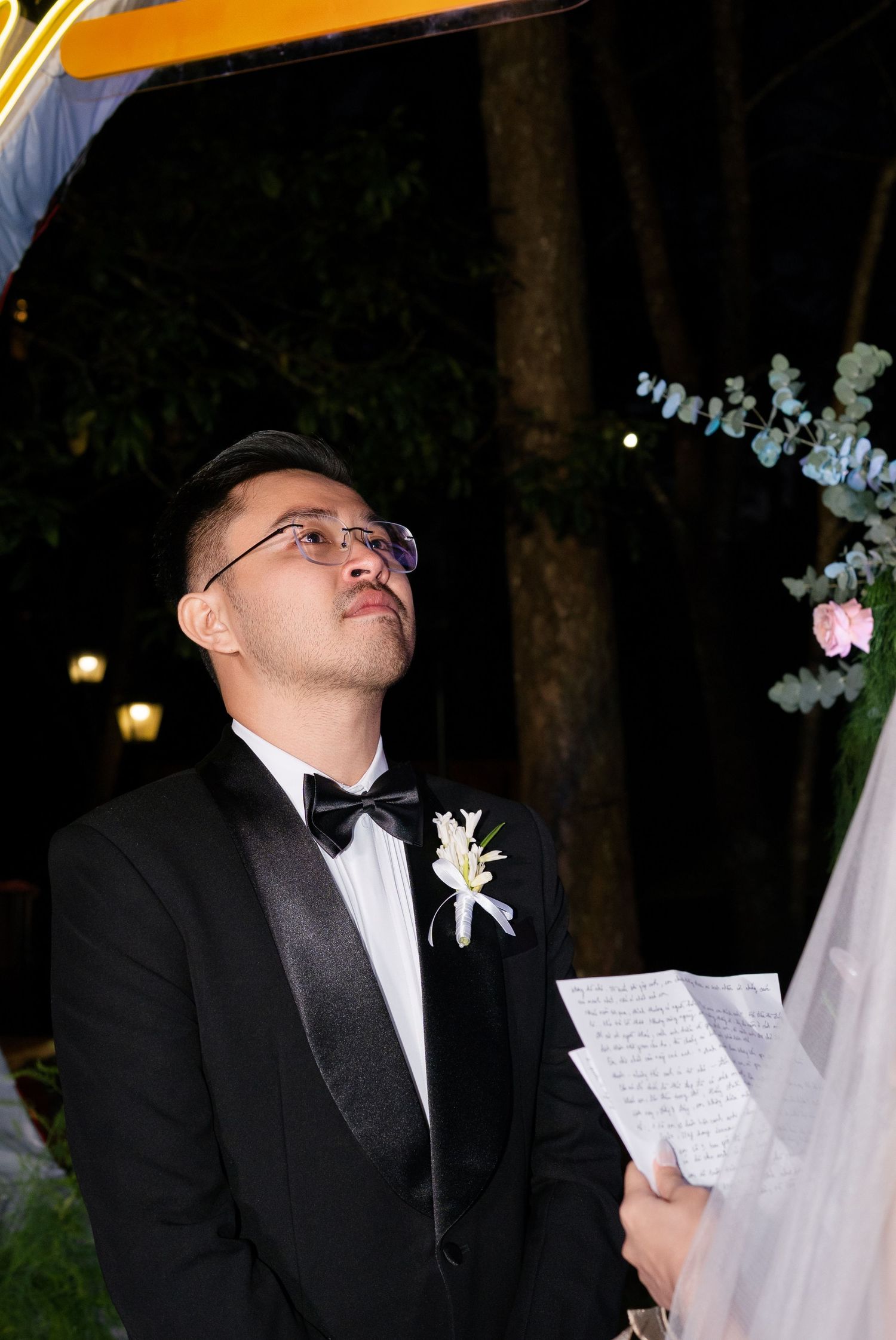 Groom in black tuxedo reads vows while looking up during elegant outdoor evening wedding ceremony.